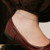 Model wearing an 18k gold-plated Tennis Anklet featuring a delicate continuous strand of clear pavé crystals around the ankle.