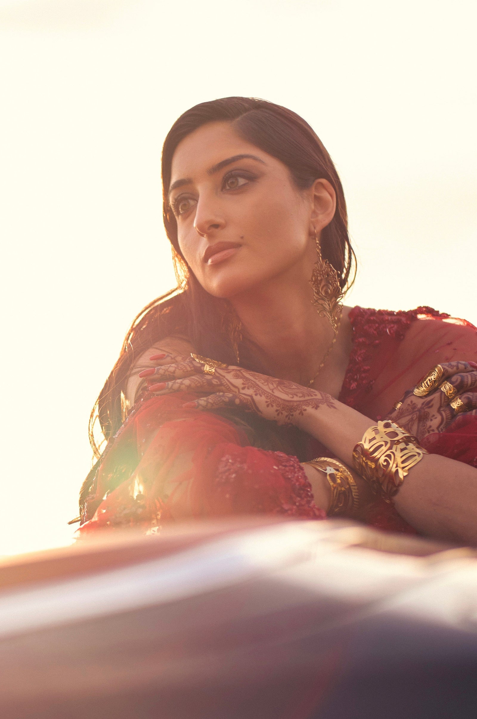 A person with long, dark hair gazes upwards and to the left. They are wearing a red, semi-sheer garment, ornate gold earrings, a gold necklace, and several gold rings. A wide, intricately cut-out gold cuff bracelet, identified as the "GRACE (Cuff)" from the Aadil x Nominal collection, adorns their wrist. Their hands are also decorated with henna patterns. The background is a soft gradient of light and warm tones, suggesting an outdoor setting with natural light.