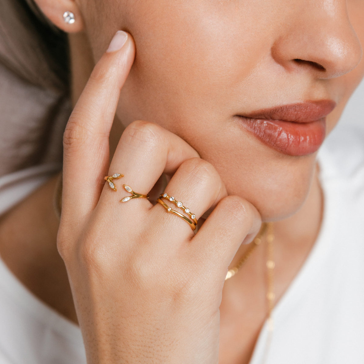 Model wearing 18k gold-plated jewelry featuring an Olive Branch Marquise Bezel Ring on the middle finger and a Five-Stone Bezel Ring stacked above a Single Bezel ring on the ring finger, paired with a Bezel Stud earring.