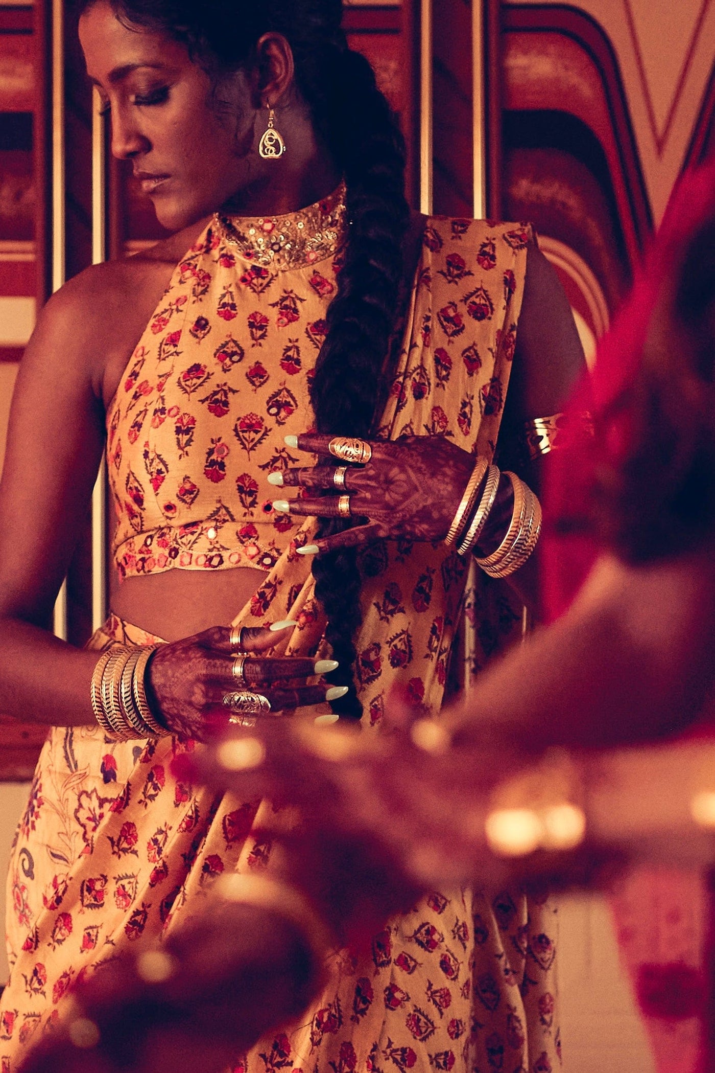 A person with dark hair styled in a long braid is adorned in traditional South Asian jewelry and clothing. They wear a patterned yellow and red sari, multiple silver bangles on their wrists, a statement gold choker, and ornate earrings. Their hands are decorated with intricate henna patterns, and they wear several rings, including a large silver ring with cut-out designs. 