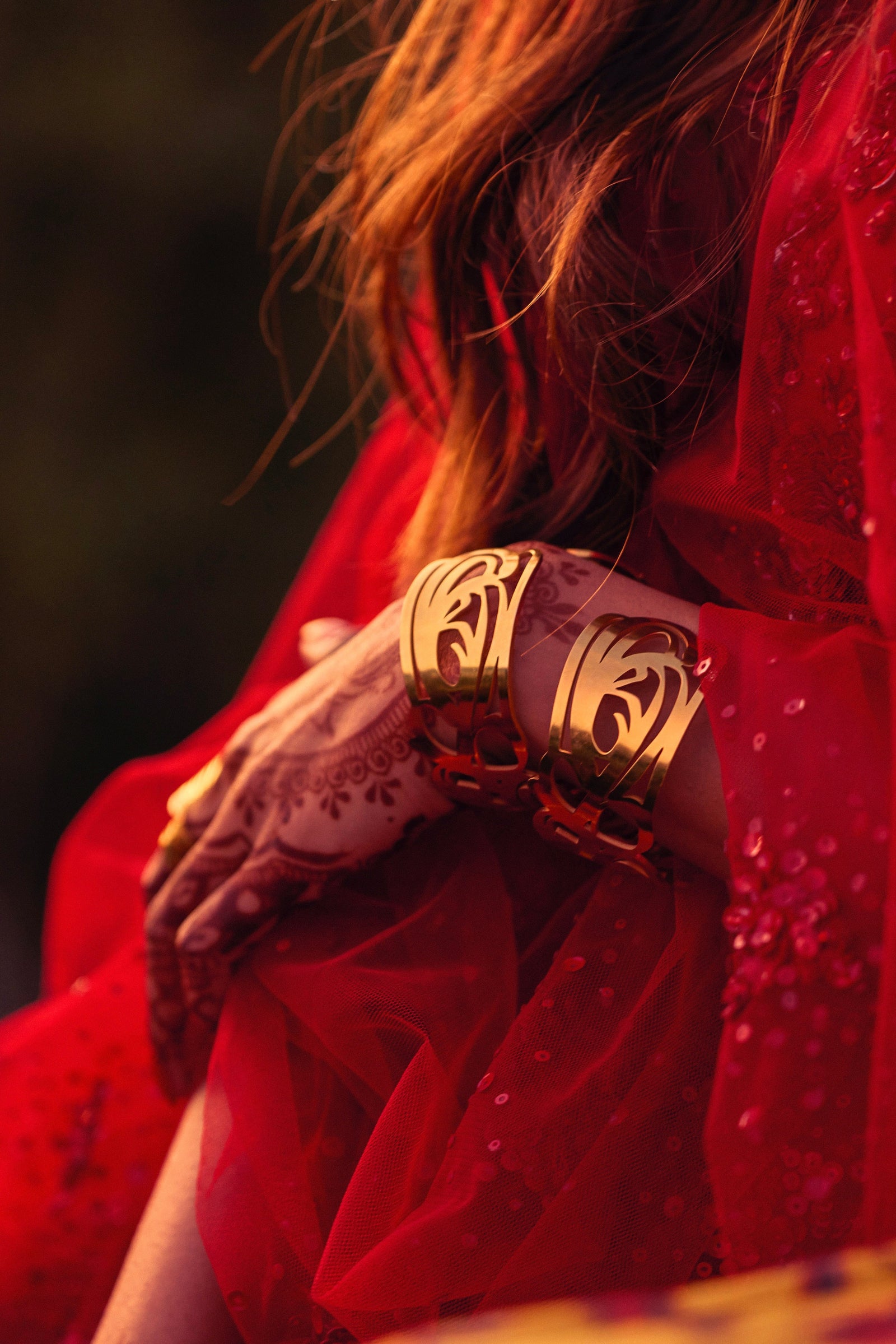 The image shows a person's hands and arms adorned with jewelry. The person is wearing a red garment. The focus is on a wide, gold cuff bracelet with intricate, swirling cut-out designs, identified as the "GRACE (Cuff)" from the Aadil x Nominal collection. The delicate patterns on the cuff create a lacy or filigree effect. The person is also wearing a gold ring and has henna designs on their hands
