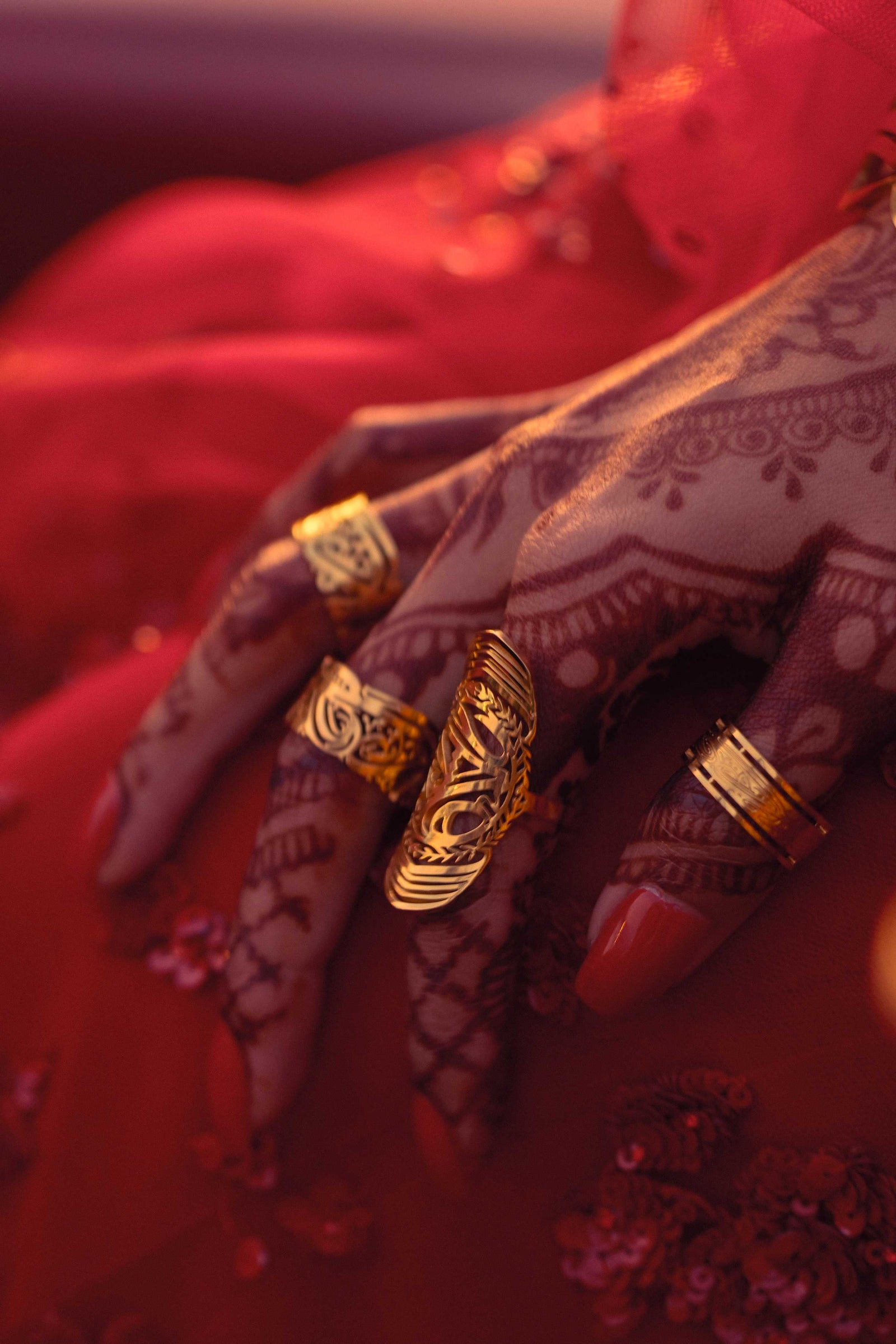 Hand decorated with henna wearing multiple gold statement rings, including the Sabireen ring with Arabic calligraphy, set against a rich red background.