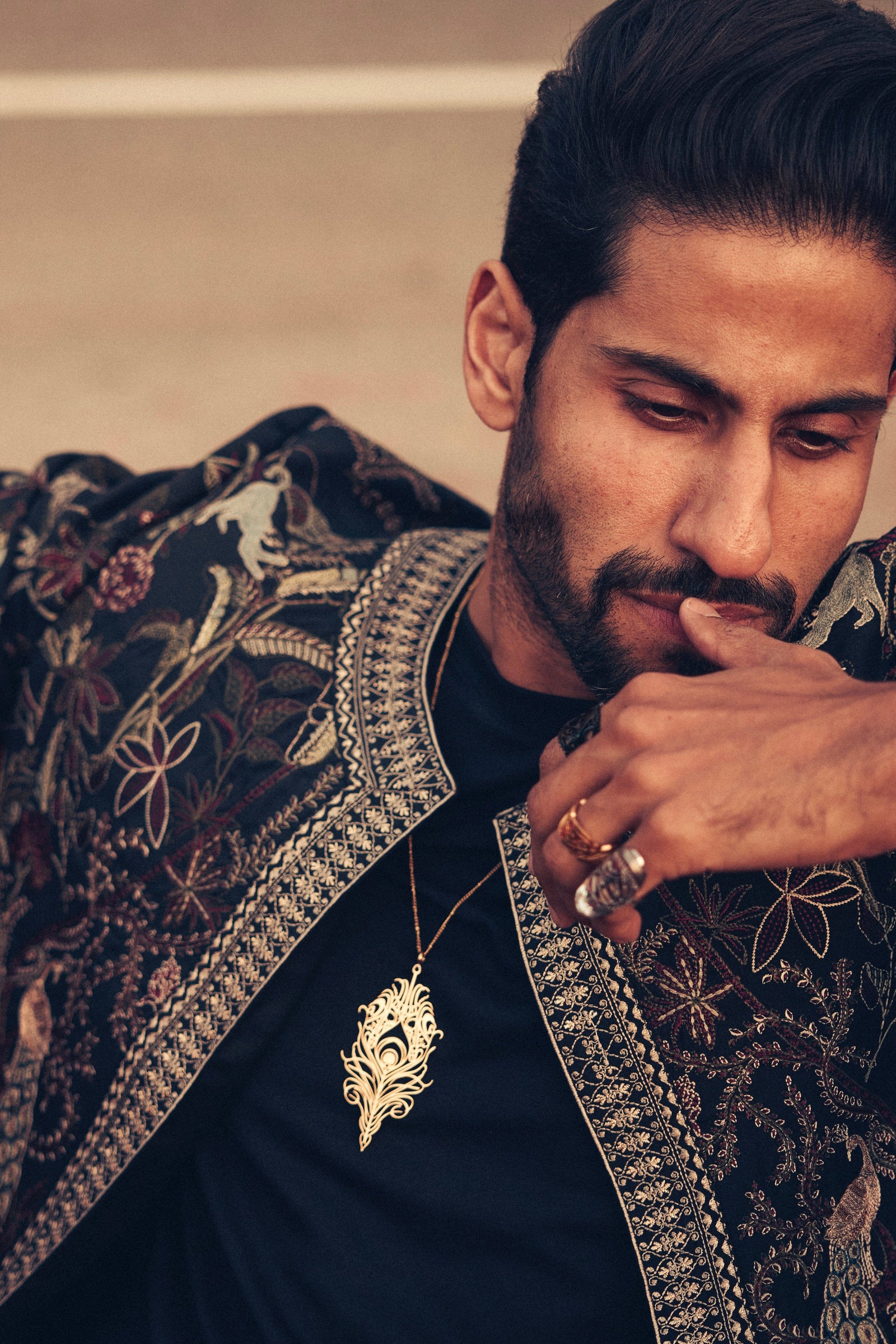 A person is wearing a delicate gold-plated chain necklace featuring a stylized, peacock feather-shaped pendant with intricate cut-out details. The necklace is shown against a dark patterned garment.