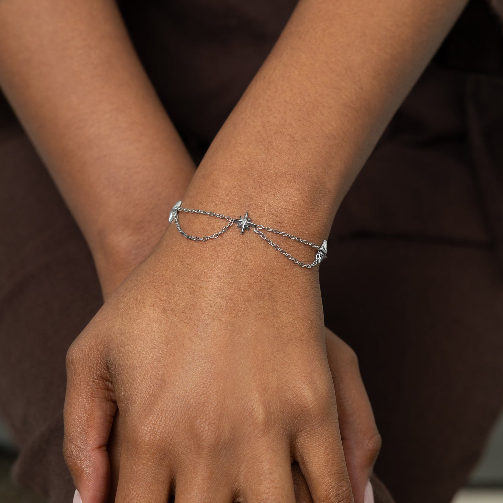 The image shows a close-up of a person’s hands resting together, with one wrist prominently displayed. They are wearing a delicate silver bracelet featuring small north star-shaped charms connected by fine chains that drape in a gentle curve across the wrist