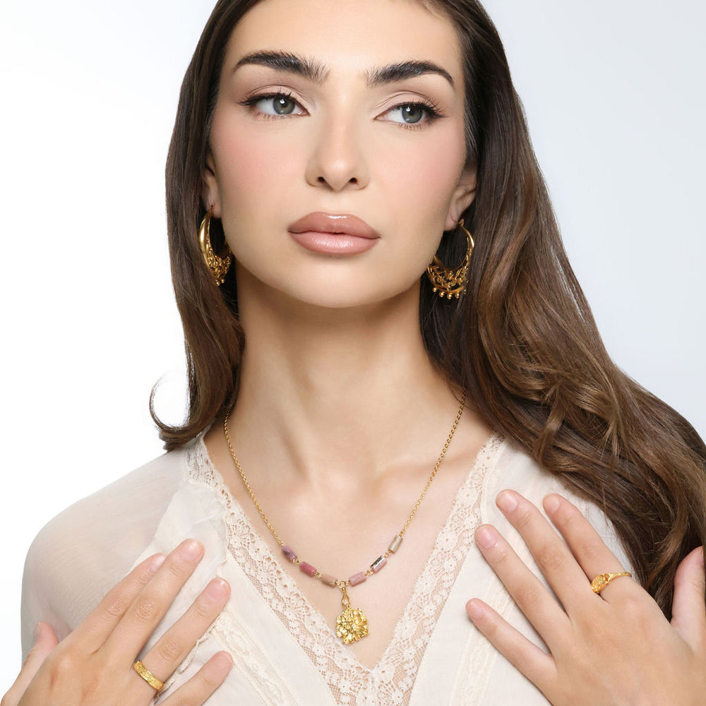 Woman wearing an 18k gold plated necklace with a textured gold flower pendant and pink and black marbled tube beads, paired with ornate gold hoop earrings
