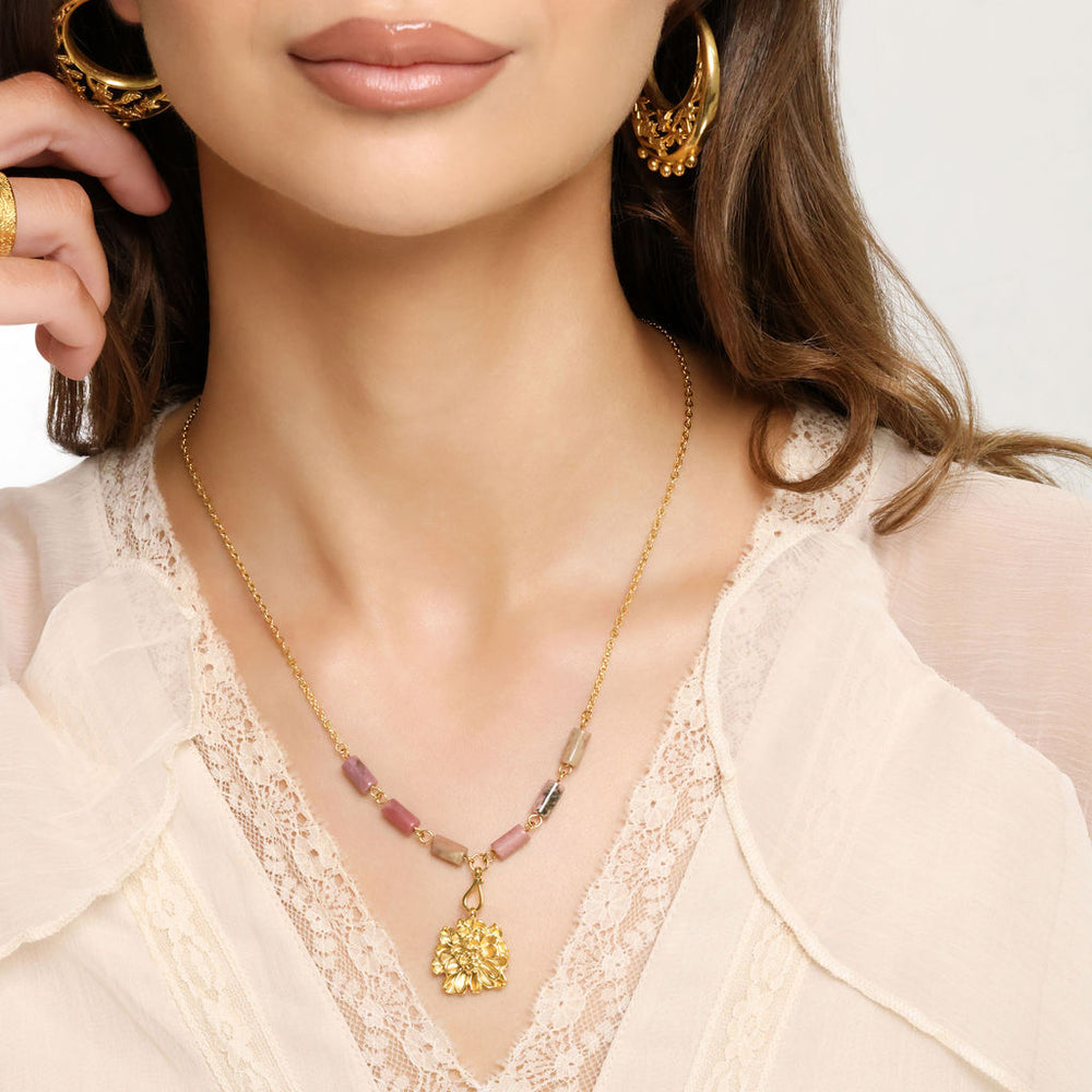 Woman wearing an 18k gold plated necklace with a textured gold flower pendant and pink and black marbled tube beads, paired with ornate gold hoop earrings