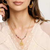 Woman wearing an 18k gold plated necklace with a textured gold flower pendant and pink and black marbled tube beads, paired with ornate gold hoop earrings