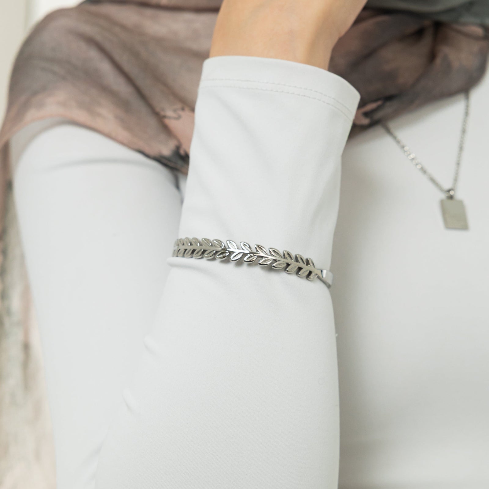 A female model with light skin tone showcasing the silver Palestine Olive Layering Bangle. The bangle drapes elegantly on her wrist, enhancing her simple attire and drawing attention to the detailed olive branch motif.