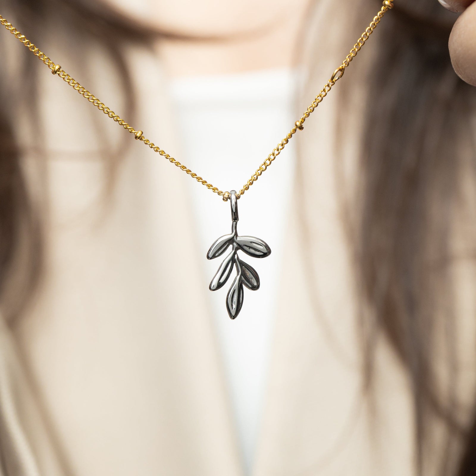 Silver olive branch charm worn on a gold beaded chain, styled against a beige outfit.