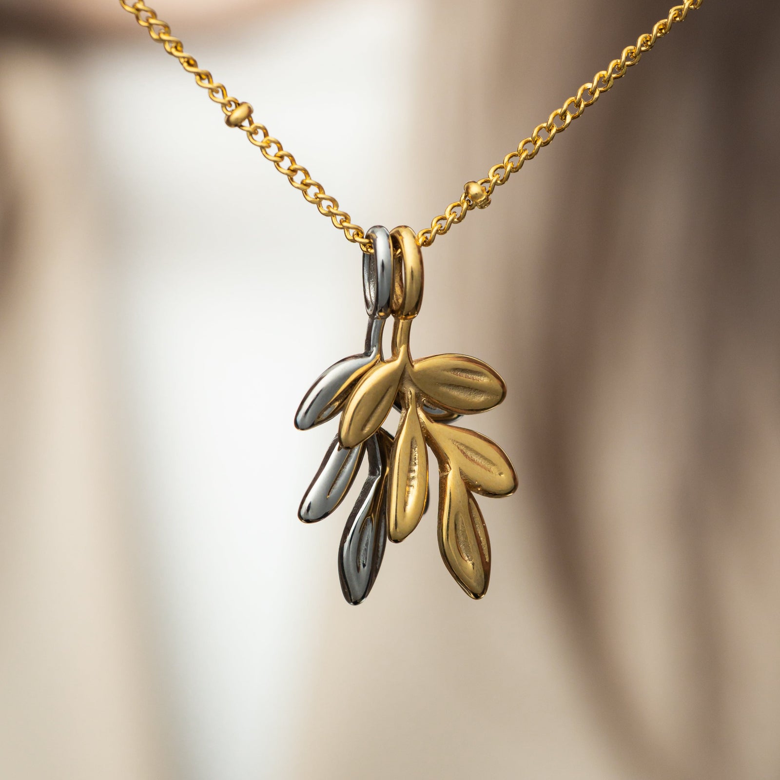 Stacked gold and silver olive branch charms layered on one beaded chain, each featuring five engraved leaves, photographed against a soft background.