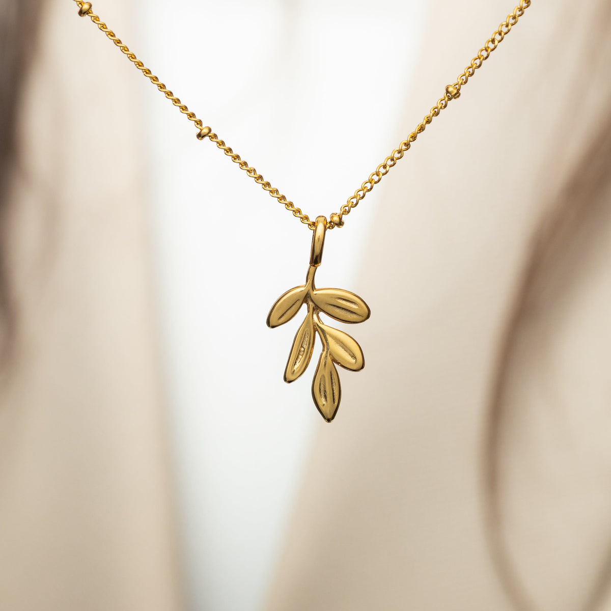 Gold olive branch charm with engraved leaves hanging from a gold beaded chain, photographed with a neutral fabric backdrop.