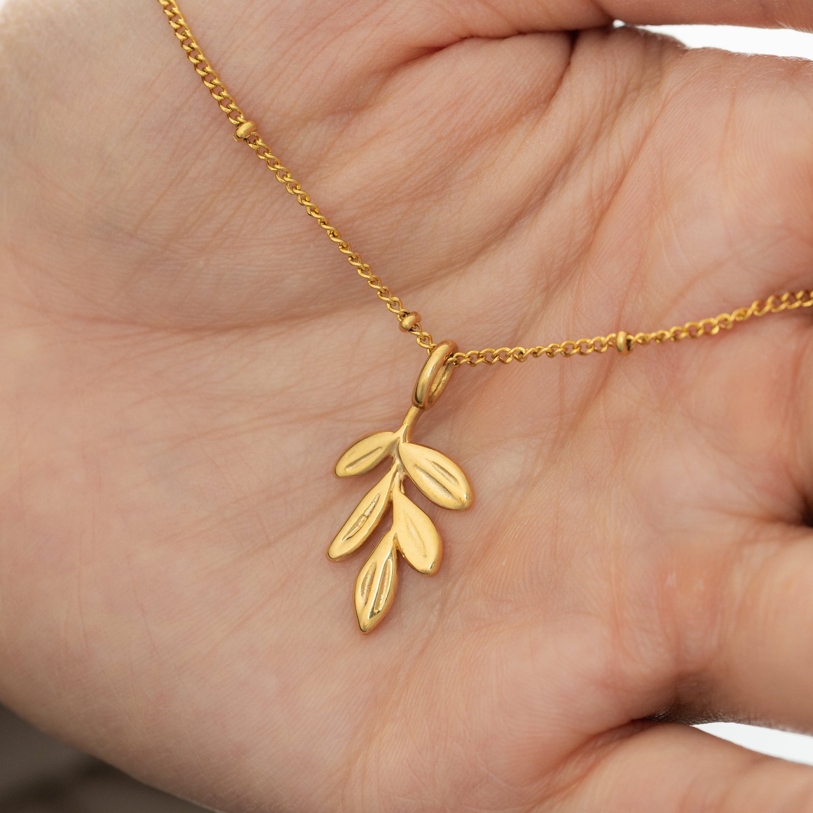 Gold olive branch charm on a beaded chain displayed resting on an open palm, showcasing size and engraved leaf details.