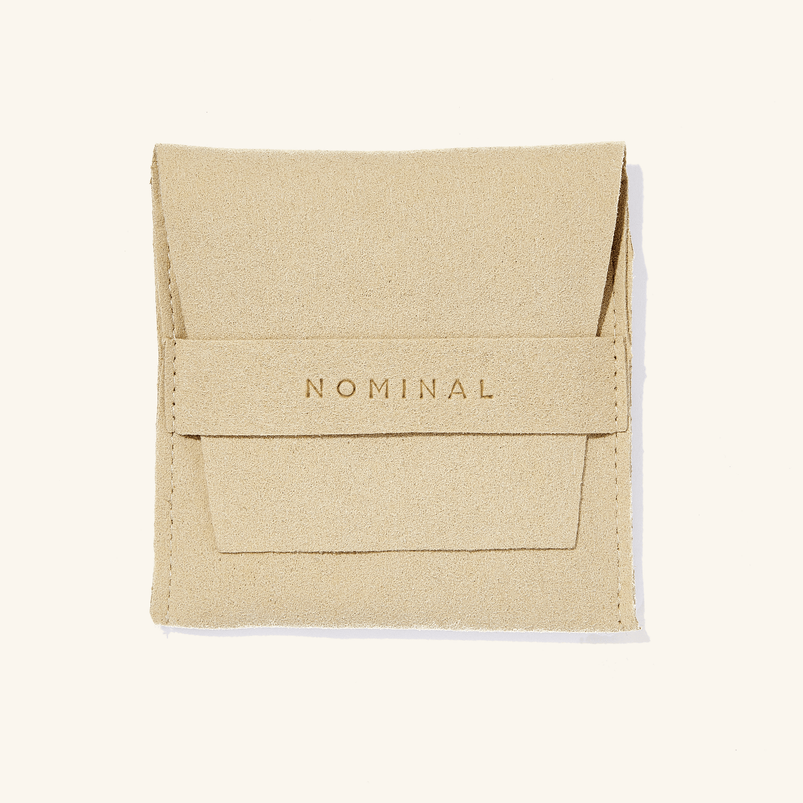 Beige suede jewelry pouch with 'NOMINAL' pressed in lettering on the front flap strap, flat against a light background.