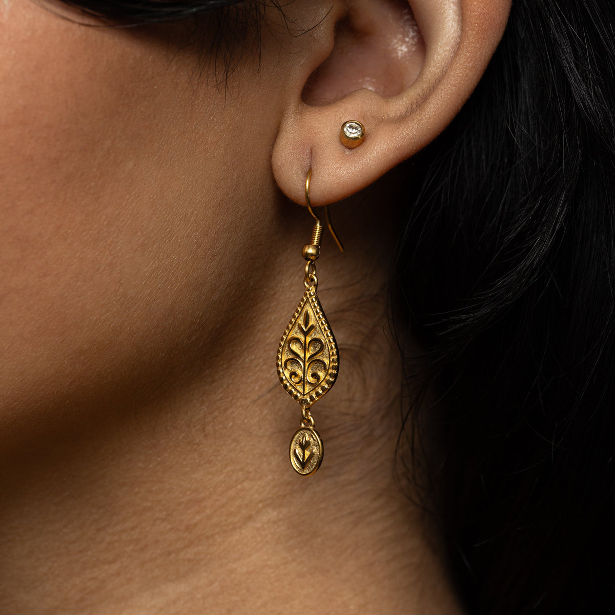 Close up photo of the gold dangling earrings worn by the model on their left ear. The model is also wearing a simple circle stud earring.