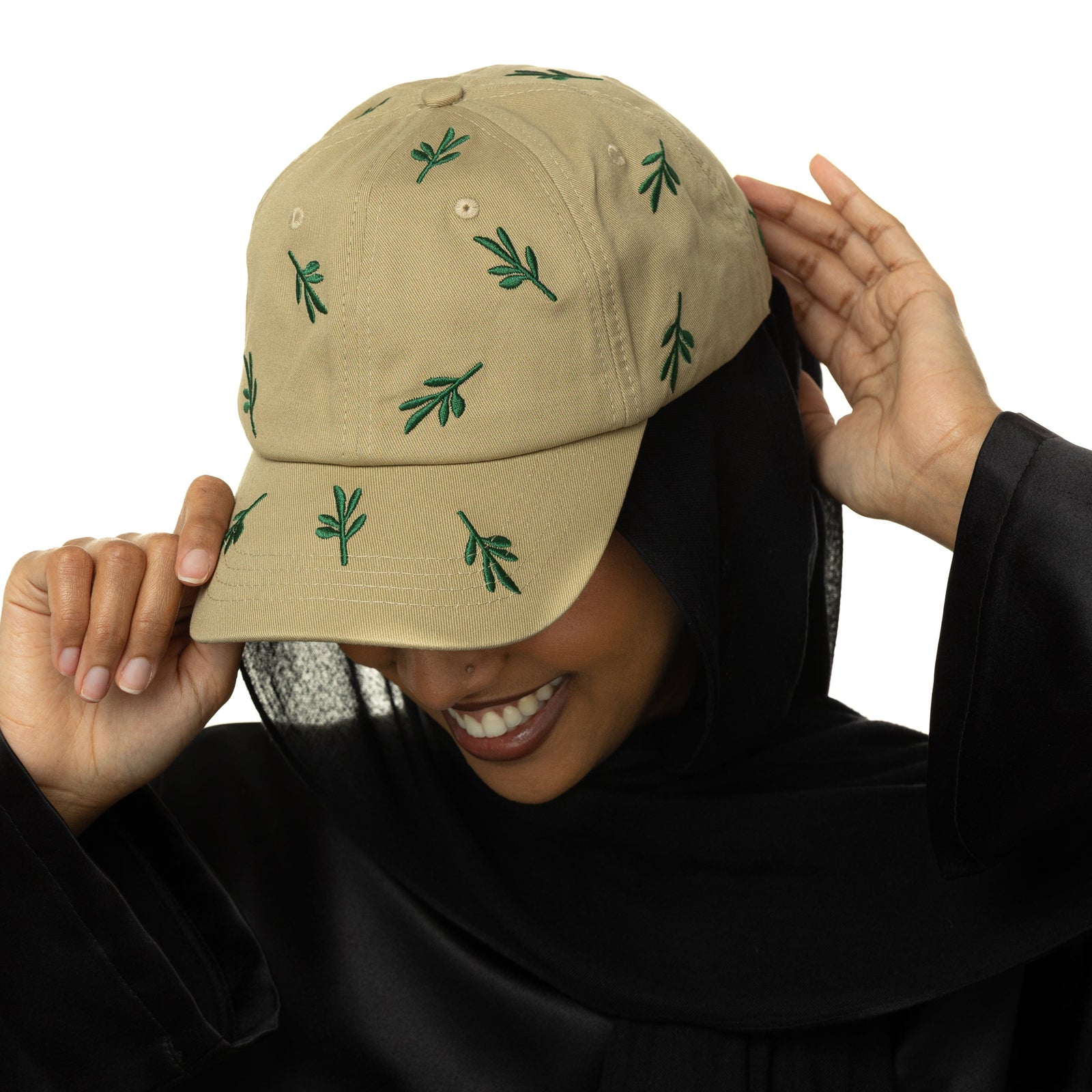 Photo of the model wearing the beige Palestinian Olive Branch with green olive branch embroidery. The model is holding the cap on their head while looking down and smiling.