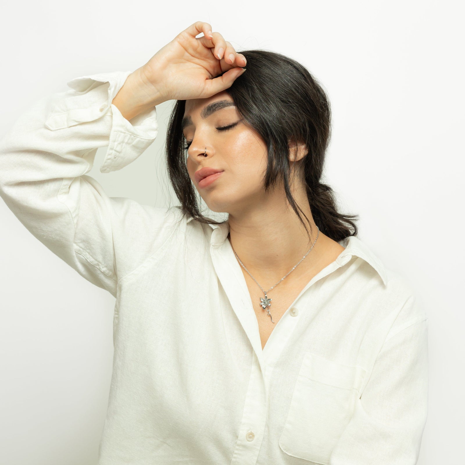 Photo of the model with her eyes are closed, and she rests one hand gently on her forehead, creating a calm and reflective pose. Her makeup appears natural and glowing, with highlighted cheekbones, defined brows, and soft lipstick. She is wearing layered necklaces, including the silver Passion' Lily Necklace.