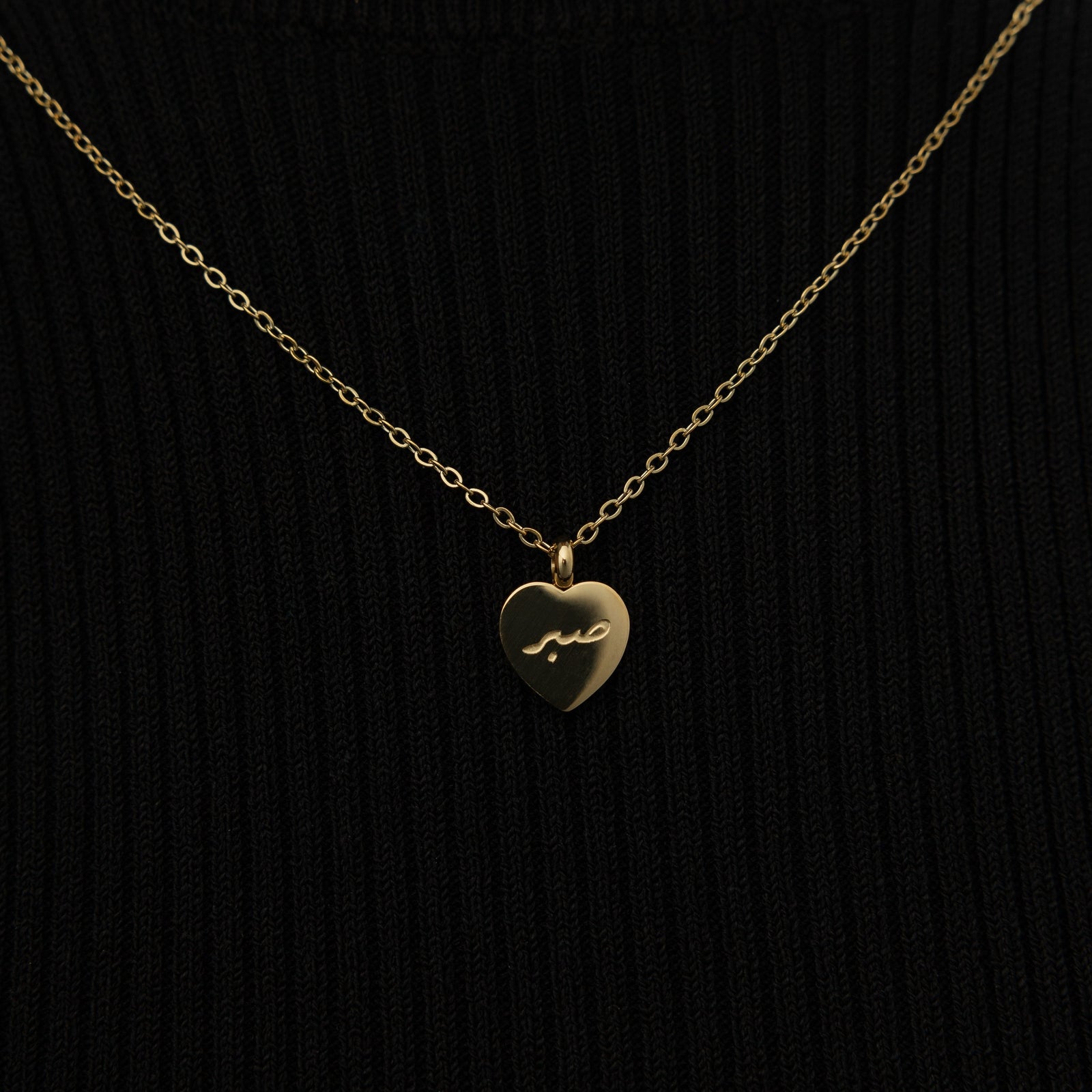 Close up of the gold Patience | صبر Charm Necklace against the black fabric of the model's clothing.