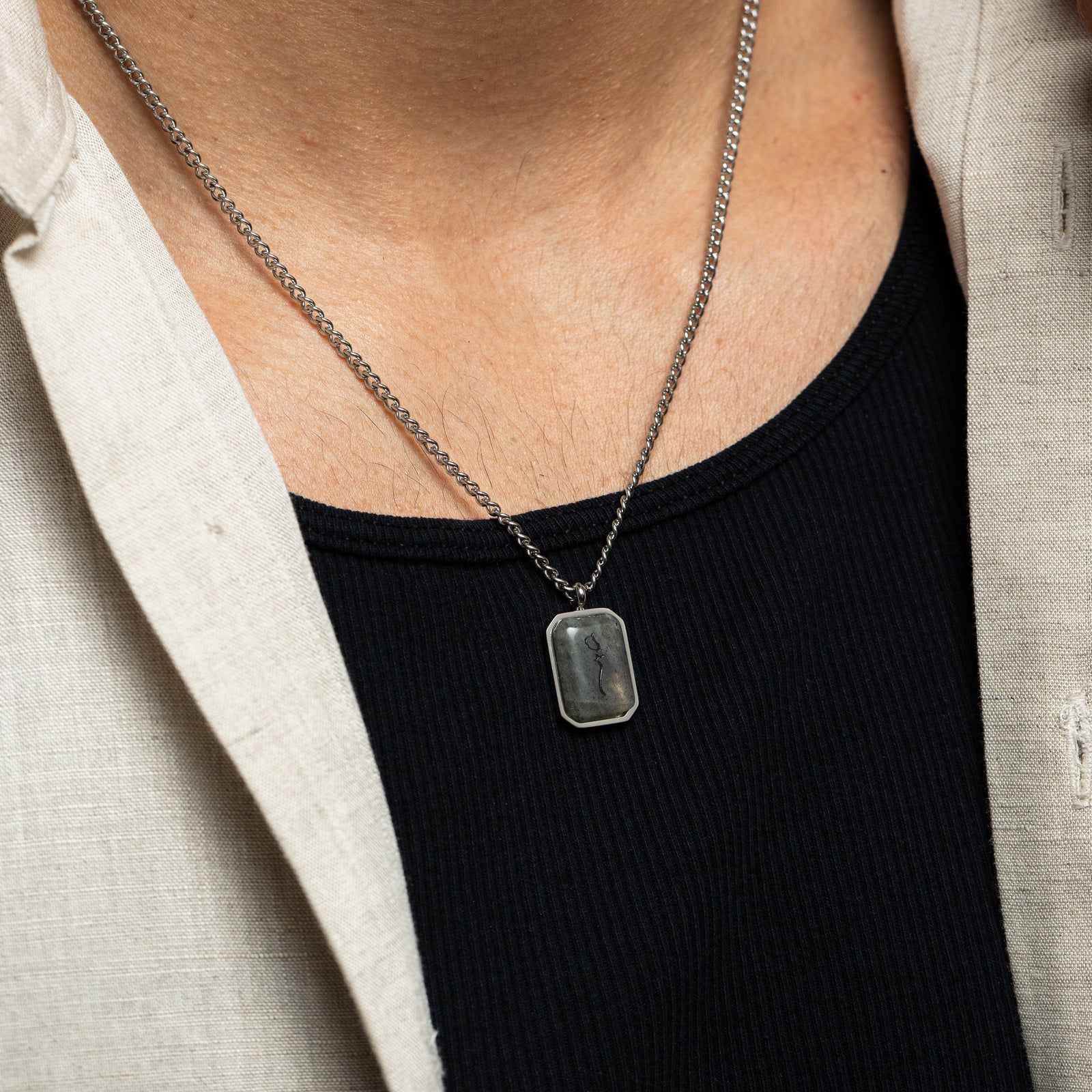 Close-up of a man wearing a silver chain necklace with a rectangular labradorite stone pendant engraved in Arabic calligraphy reading "Sabr," styled over a black ribbed shirt with beige shirt edges visible.