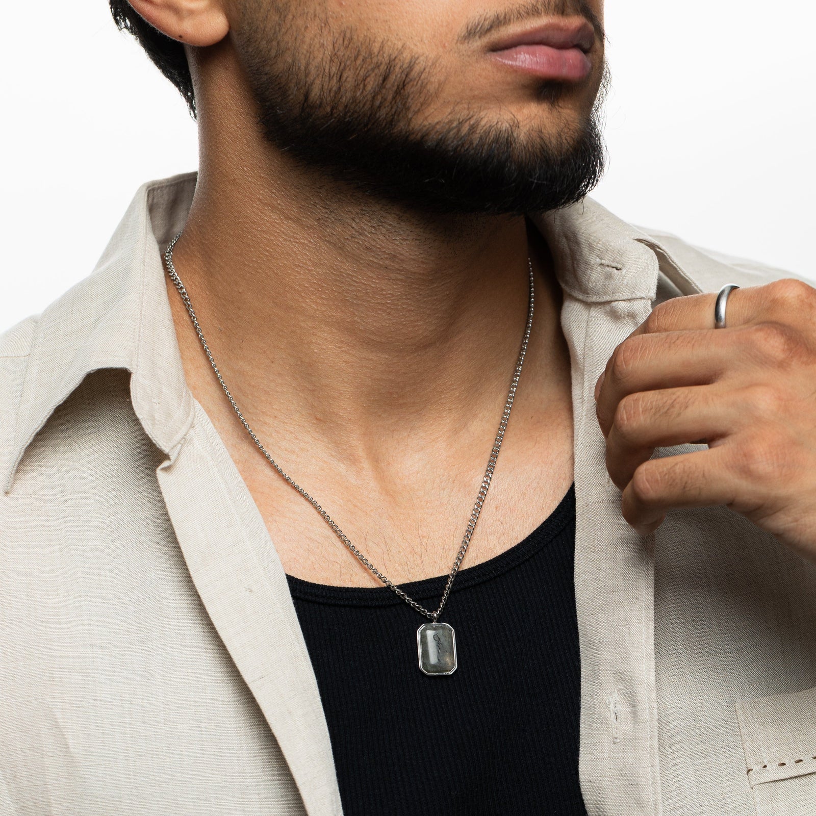 Close-up of a man wearing a silver chain necklace with a rectangular labradorite stone pendant engraved with "Sabr" in Arabic calligraphy, styled with a black ribbed shirt and beige open shirt, hand pulling shirt collar slightly.