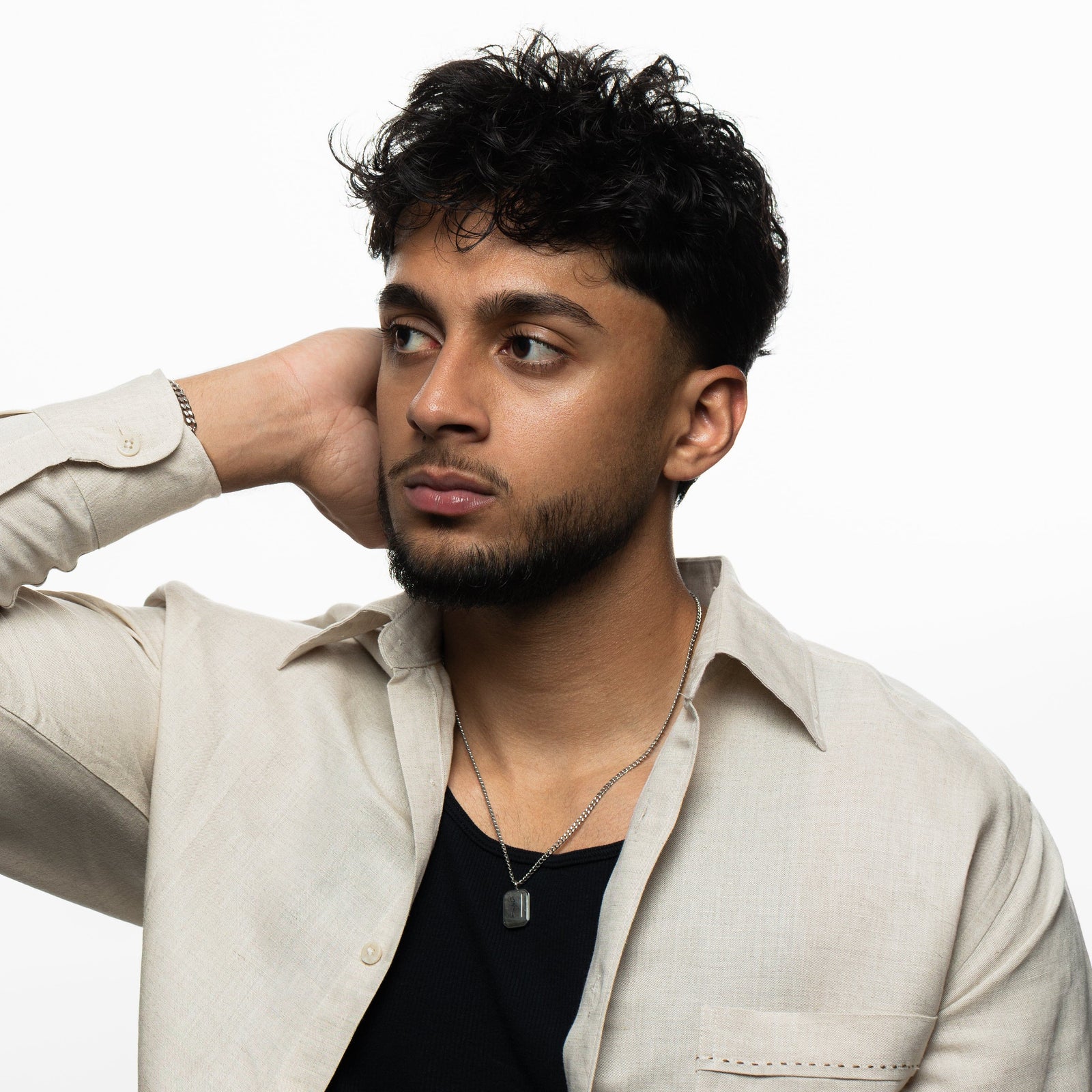 Man wearing a silver chain necklace with a rectangular labradorite stone pendant engraved with "Sabr" in Arabic calligraphy, styled with a black ribbed shirt and beige open shirt, hand resting on head.