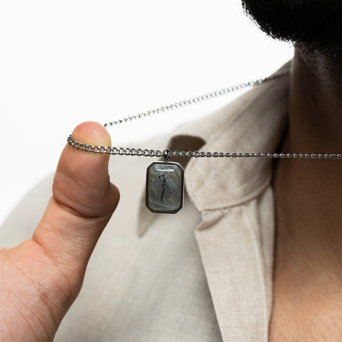 Man holding a silver chain necklace with a rectangular labradorite pendant engraved with "Sabr" in Arabic calligraphy, styled against a beige shirt.