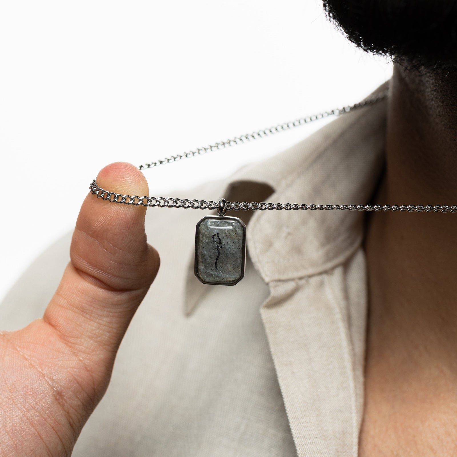 Man holding a silver chain necklace with a rectangular labradorite pendant engraved with "Sabr" in Arabic calligraphy, styled against a beige shirt.