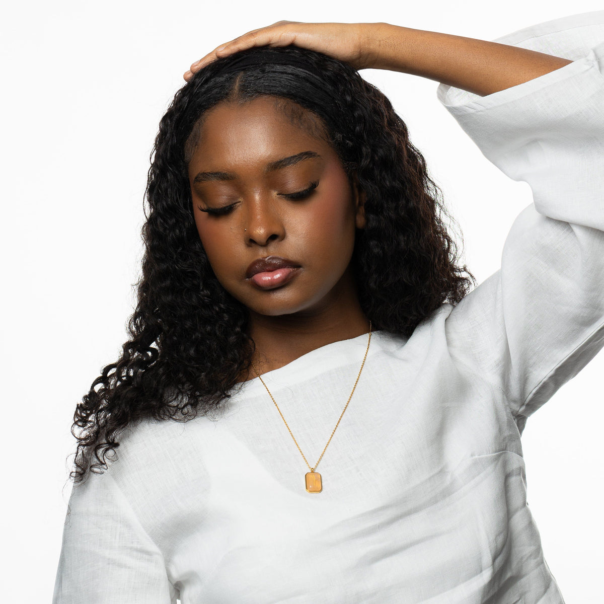 Woman wearing a gold chain necklace with a rectangular rose quartz pendant engraved with "Sabr" in Arabic calligraphy, styled with a white blouse, hand resting above her head.