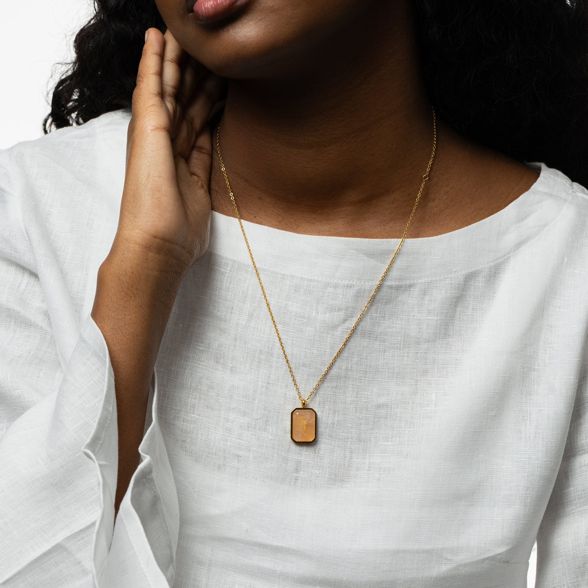 Close-up of a woman wearing a gold chain necklace with a rectangular rose quartz pendant engraved with "Sabr" in Arabic calligraphy, styled with a white blouse, hand resting gently near her neck.