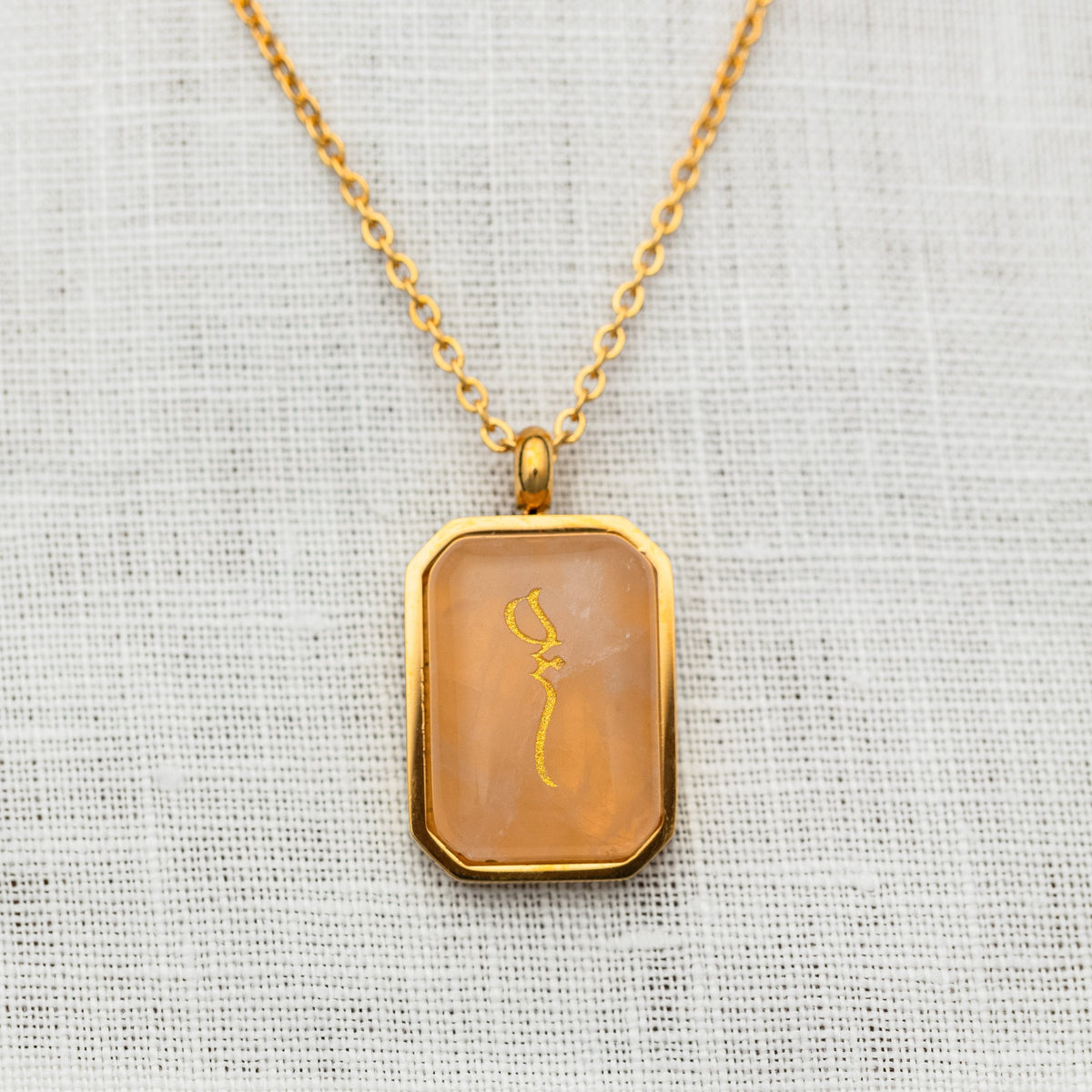 Close-up of a gold chain necklace with a rectangular rose quartz stone pendant engraved with "Sabr" in Arabic calligraphy, placed against a textured white fabric background.