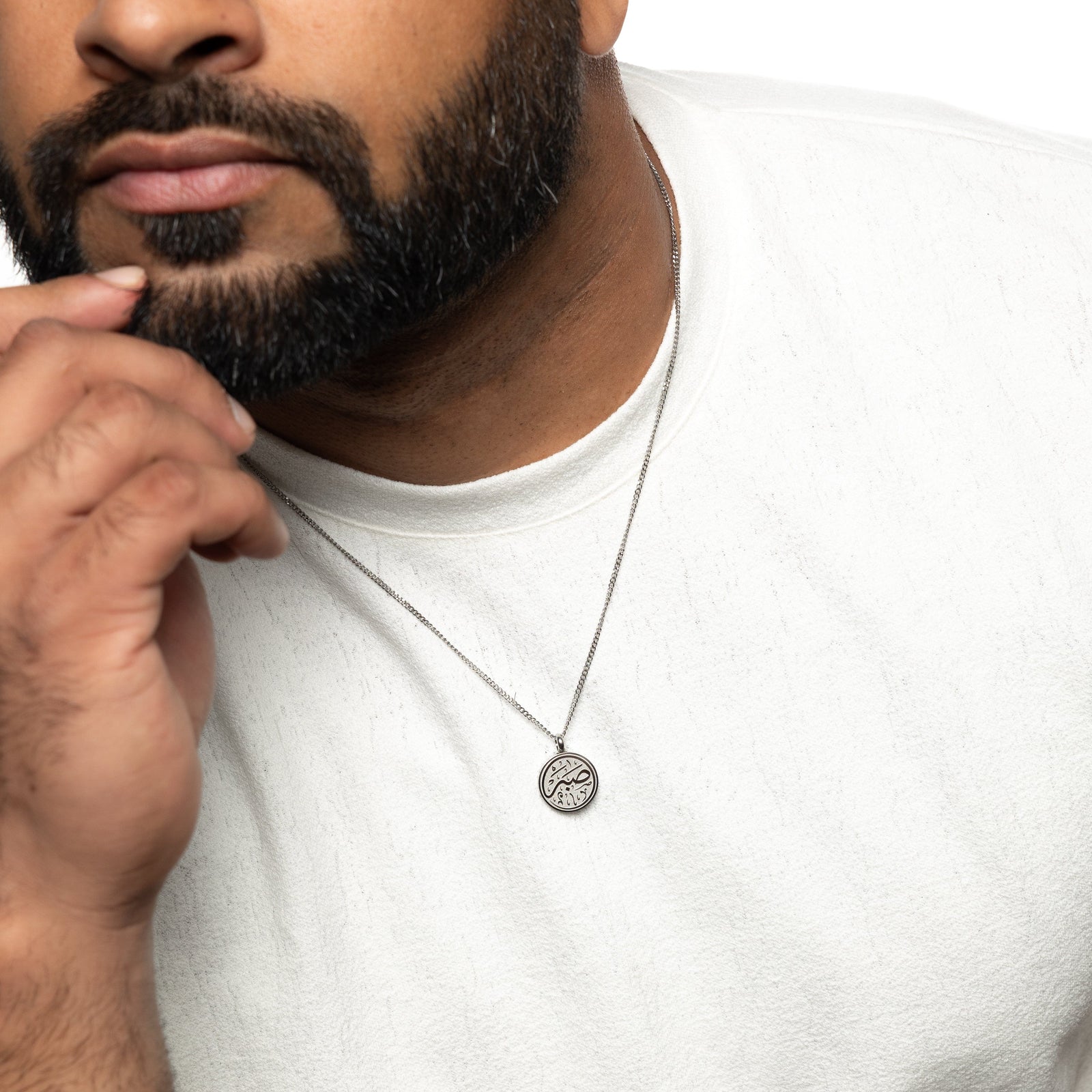 Man wearing a silver patience token necklace over a white shirt, resting his hand near his chin.