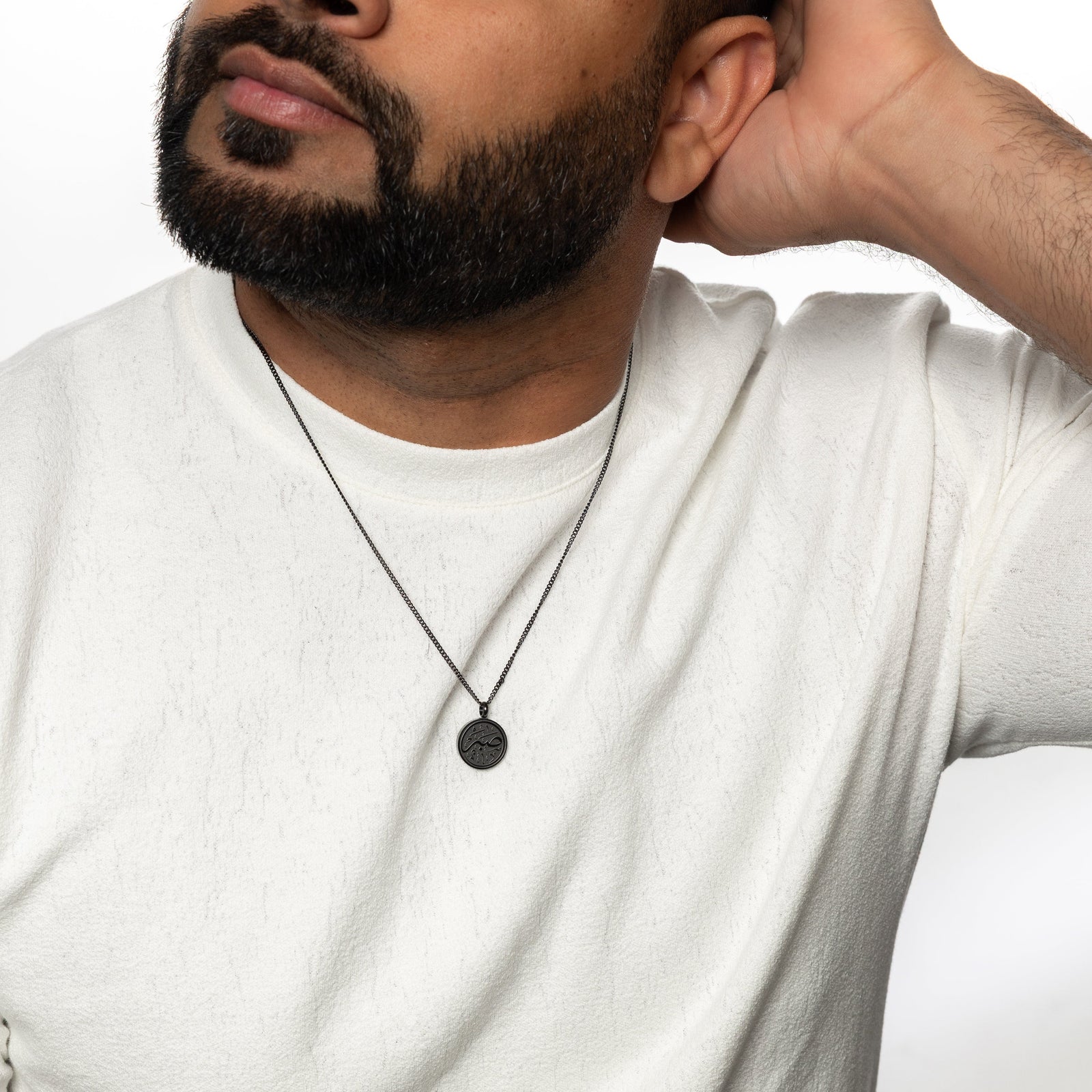Man wearing a black Patience token necklace over a white shirt, arm resting behind his head.