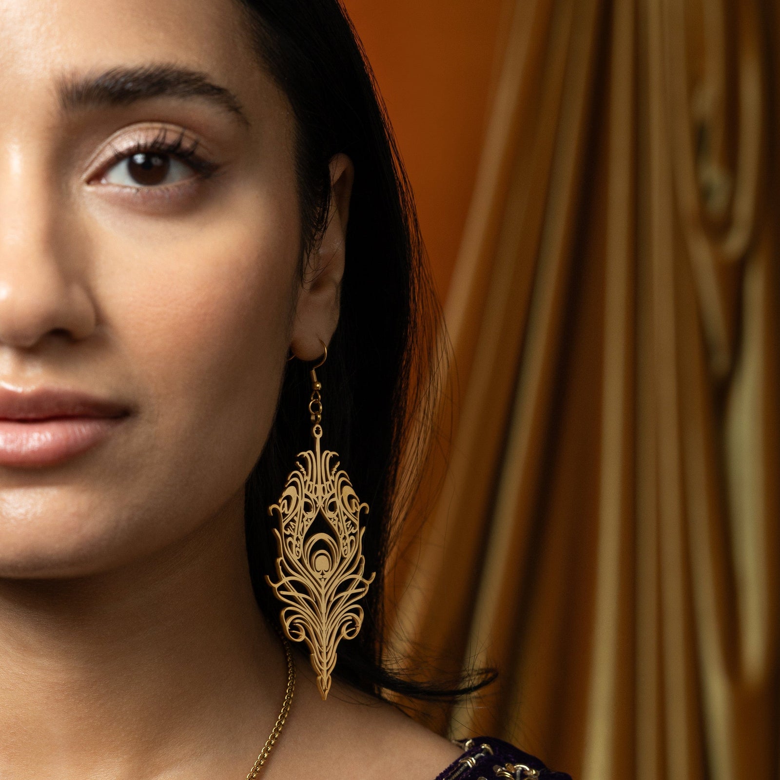 A person with long, dark, slightly wavy hair is shown in profile, facing left. They are wearing a gold "Peacock Feather Earring" from the Aadil x Nominal collection. The earring is intricately designed in the shape of a peacock feather with detailed cut-out patterns. The person is also wearing a red garment with embellishments and has henna on their hands. The background is softly blurred, suggesting an indoor setting.