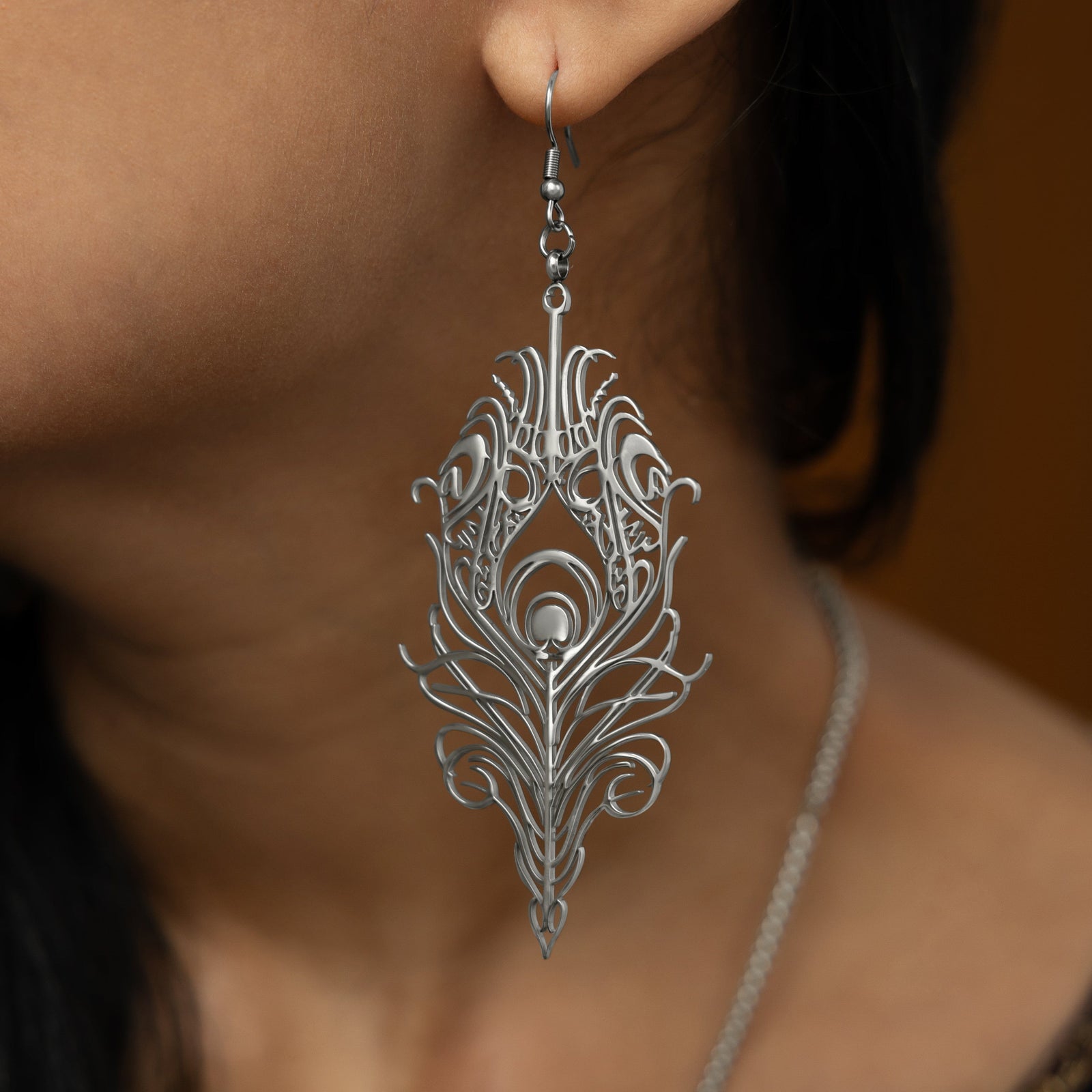 A close-up of a person's ear wearing a dangling silver earring shaped like a peacock feather with intricate cut-out details.