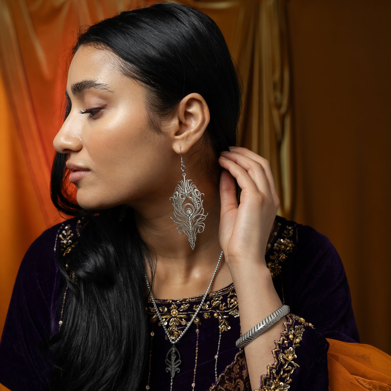 A person with dark hair is shown in profile, wearing a silver peacock feather earring, a matching silver necklace, and a silver bangle with a chevron pattern. They are wearing a dark blue velvet top with gold embroidery and have their hand near their ear. The background is a warm, draped fabric.