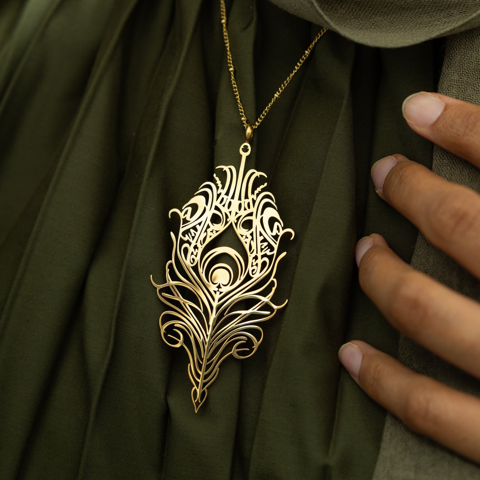 A delicate gold-plated chain necklace featuring a stylized, peacock feather-shaped pendant with intricate cut-out details. The pendant and chain are displayed against a light beige background.