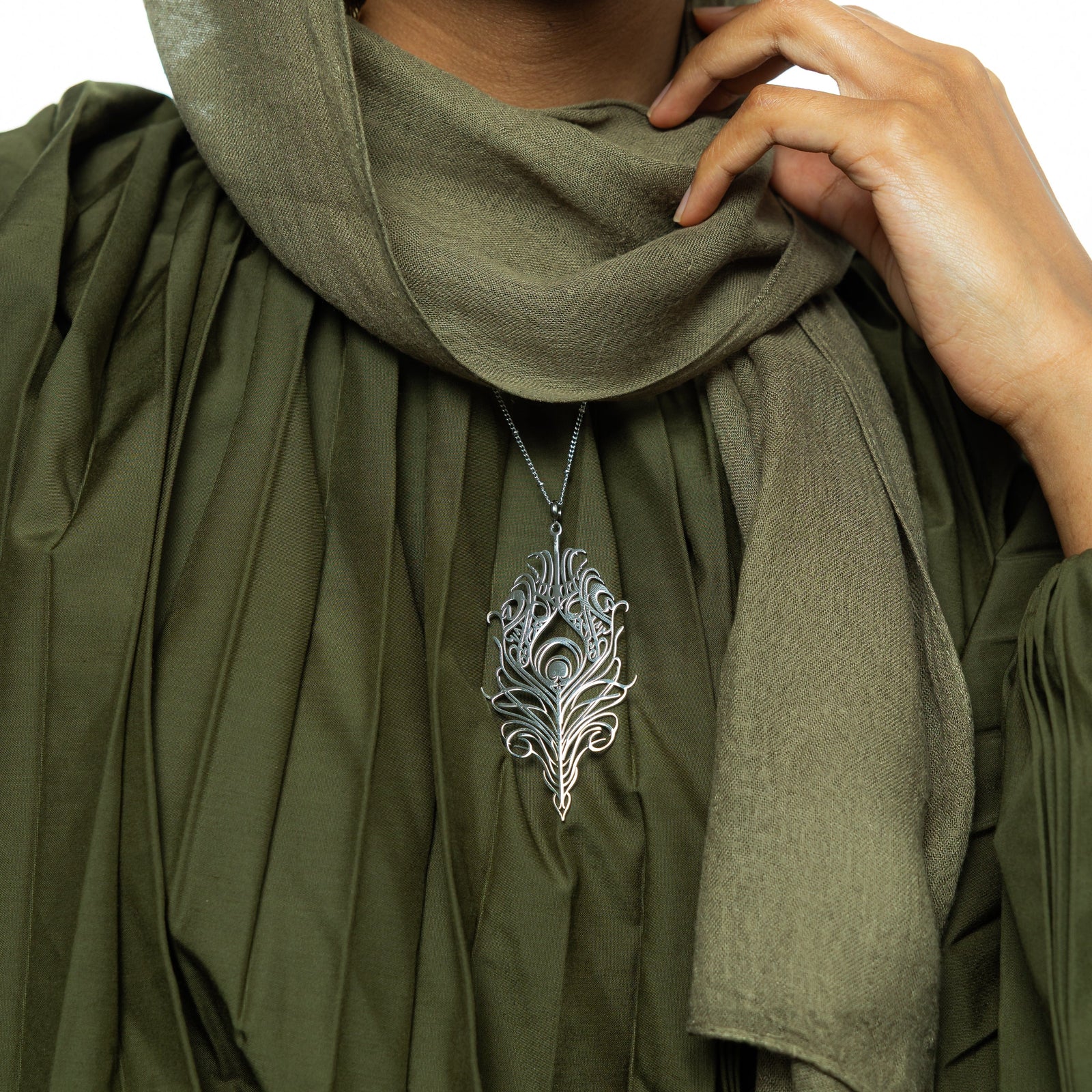 A delicate silver-plated chain necklace featuring a stylized, peacock feather-shaped pendant with intricate cut-out details. The necklace is worn by a person with dark hair and is shown against a blurred green garment. A hand with light-colored nail polish is gently holding the chain near the pendant.