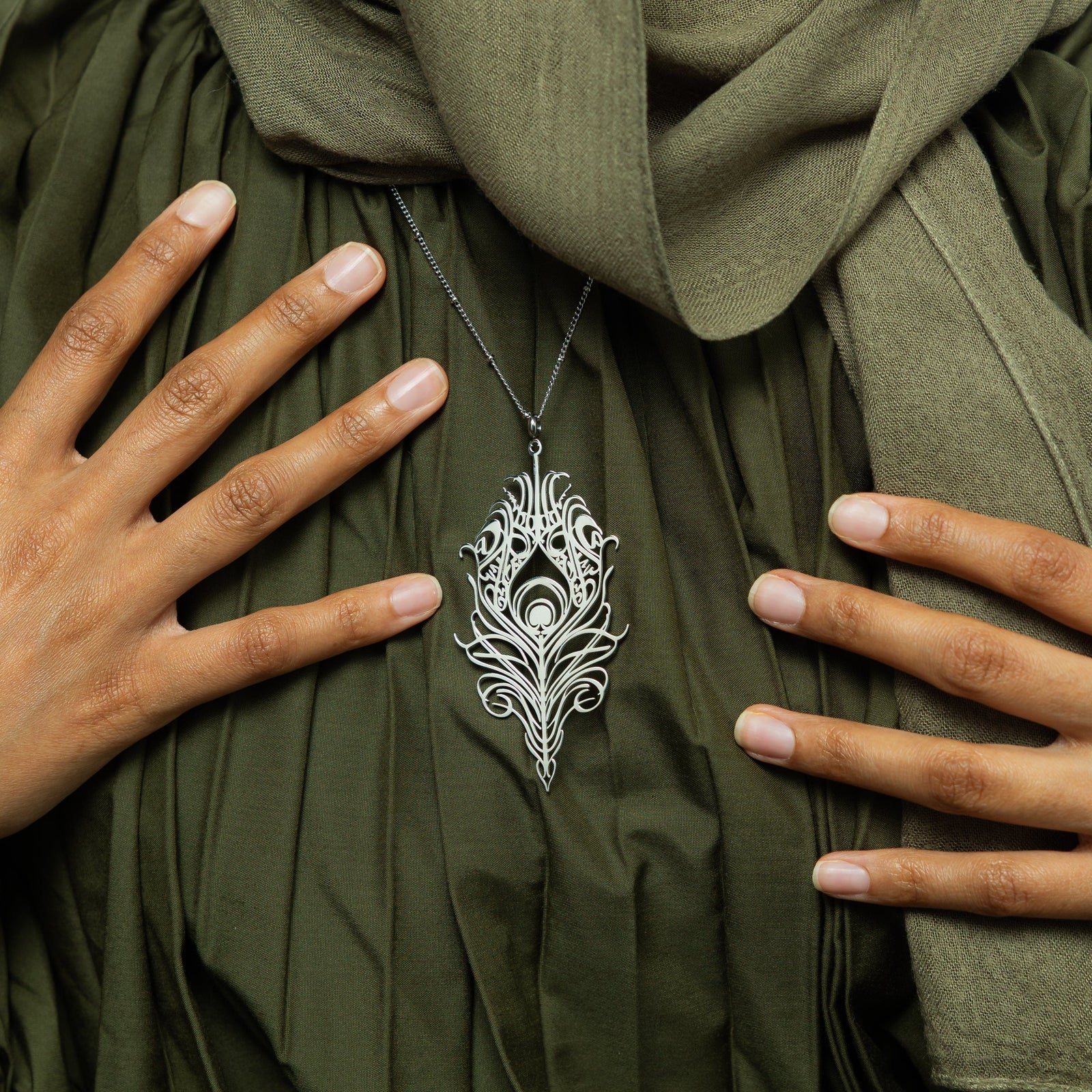 A delicate silver-plated chain necklace featuring a stylized, peacock feather-shaped pendant with intricate cut-out details. The necklace is worn against a textured green garment. Two hands with light-colored nail polish are gently placed on either side of the pendant.