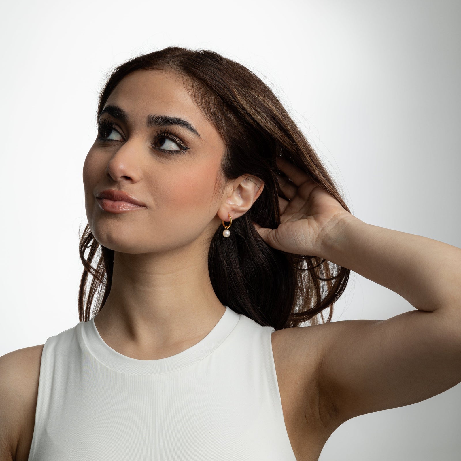 Photo of the model looking upward and holding their hair behind their left ear to show the Pearl Hoop Earrings they are wearing.