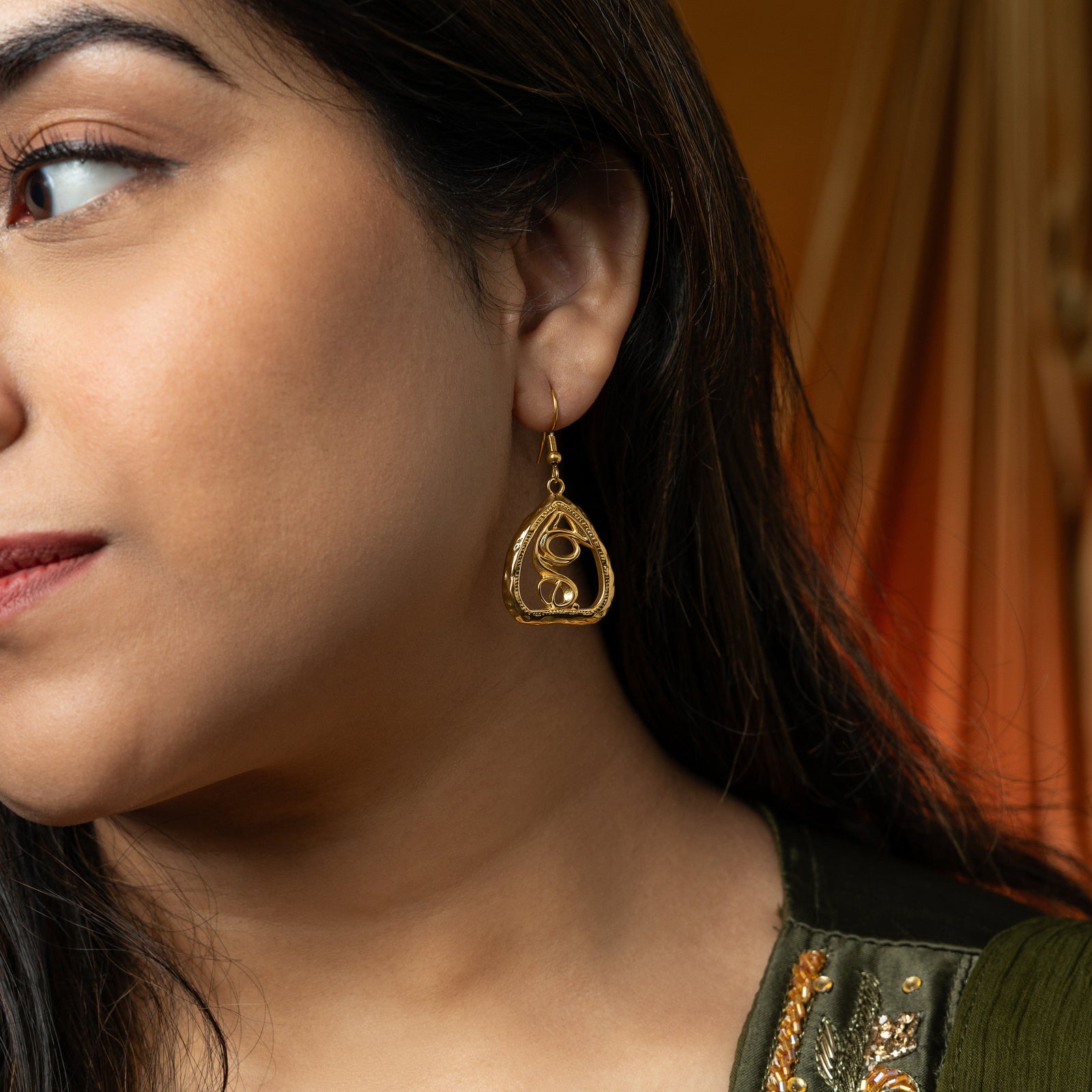 A person with long, dark hair is shown in profile, looking down and to the left. They are wearing silver "Peepal Earrings" from the Aadil x Nominal collection. The earrings have a unique, abstract design with swirling lines enclosed within a roughly triangular frame. The person is also wearing a silver necklace and a light-colored garment with mirror embellishments. Their hand, adorned with intricate henna patterns, is near their face. The background shows part of a red car interior and a blurred outdoor.
