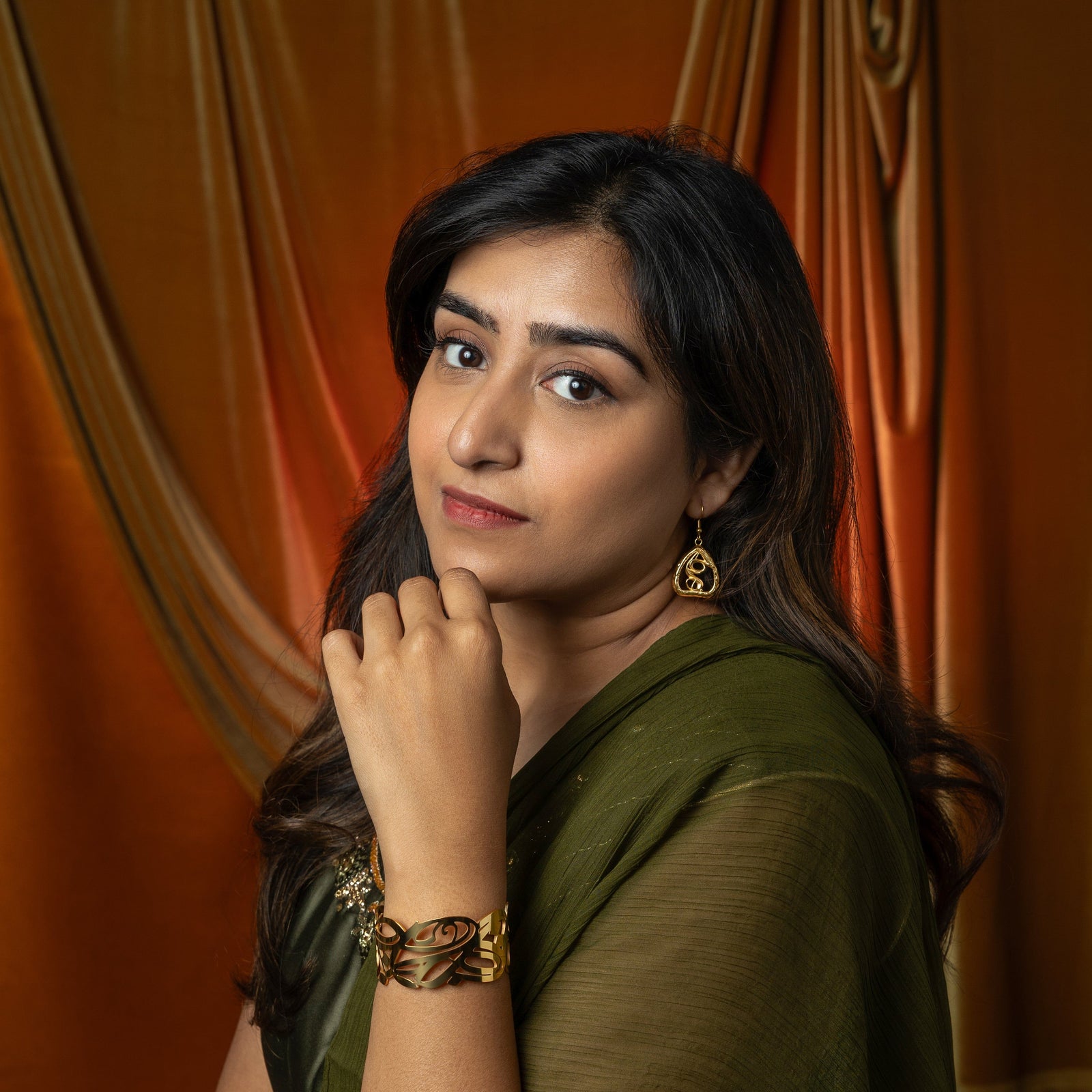 A person with long, dark hair is shown in profile, looking down and to the left. They are wearing silver "Peepal Earrings" from the Aadil x Nominal collection. The earrings have a unique, abstract design with swirling lines enclosed within a roughly triangular frame. The person is also wearing a silver necklace and a light-colored garment with mirror embellishments. Their hand, adorned with intricate henna patterns, is near their face. The background shows part of a red car interior and a blurred outdoor sc