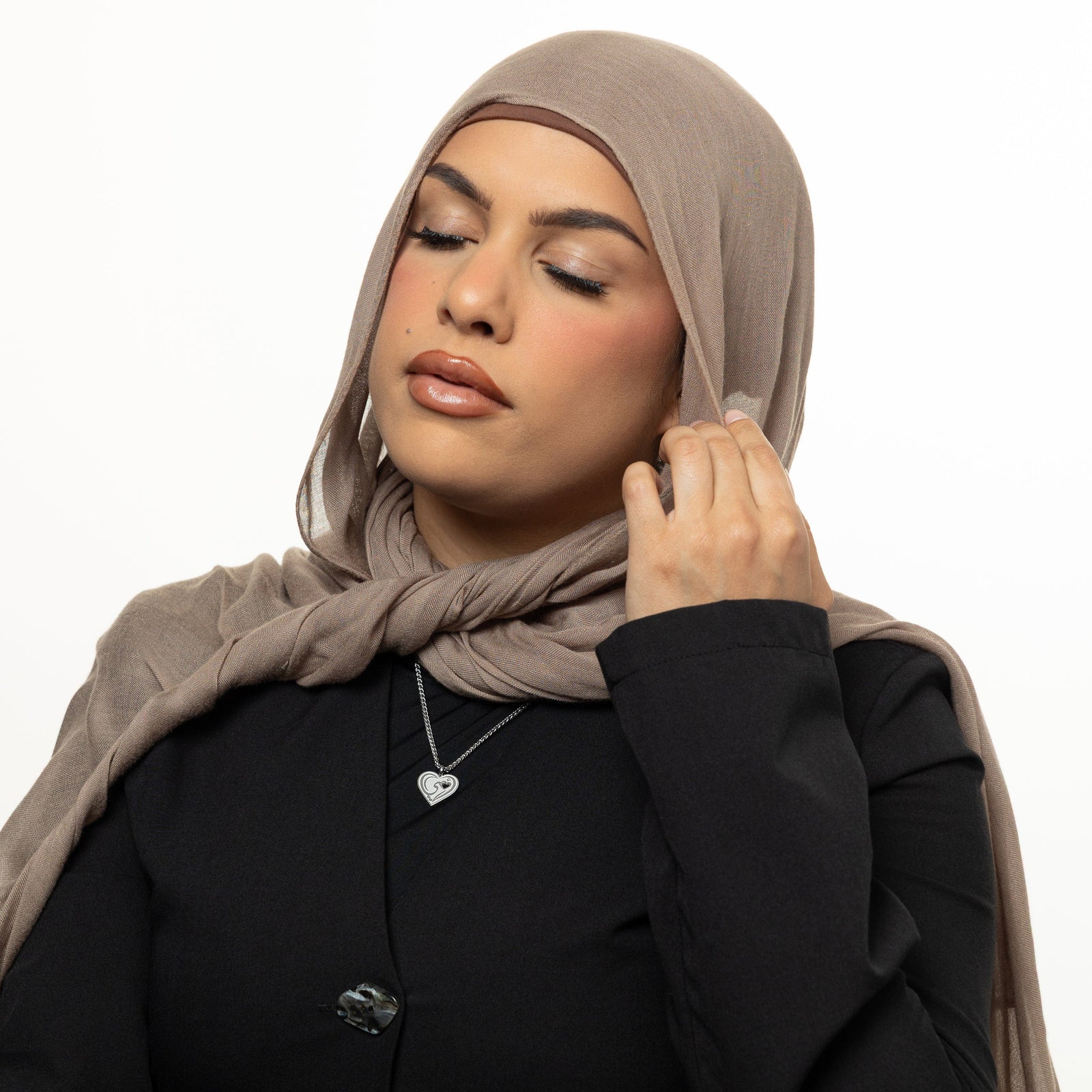 Photo of the model touching their beige hijab and their eyes closed. The model is wearing the silver "Piece of Me" Mama Necklace.