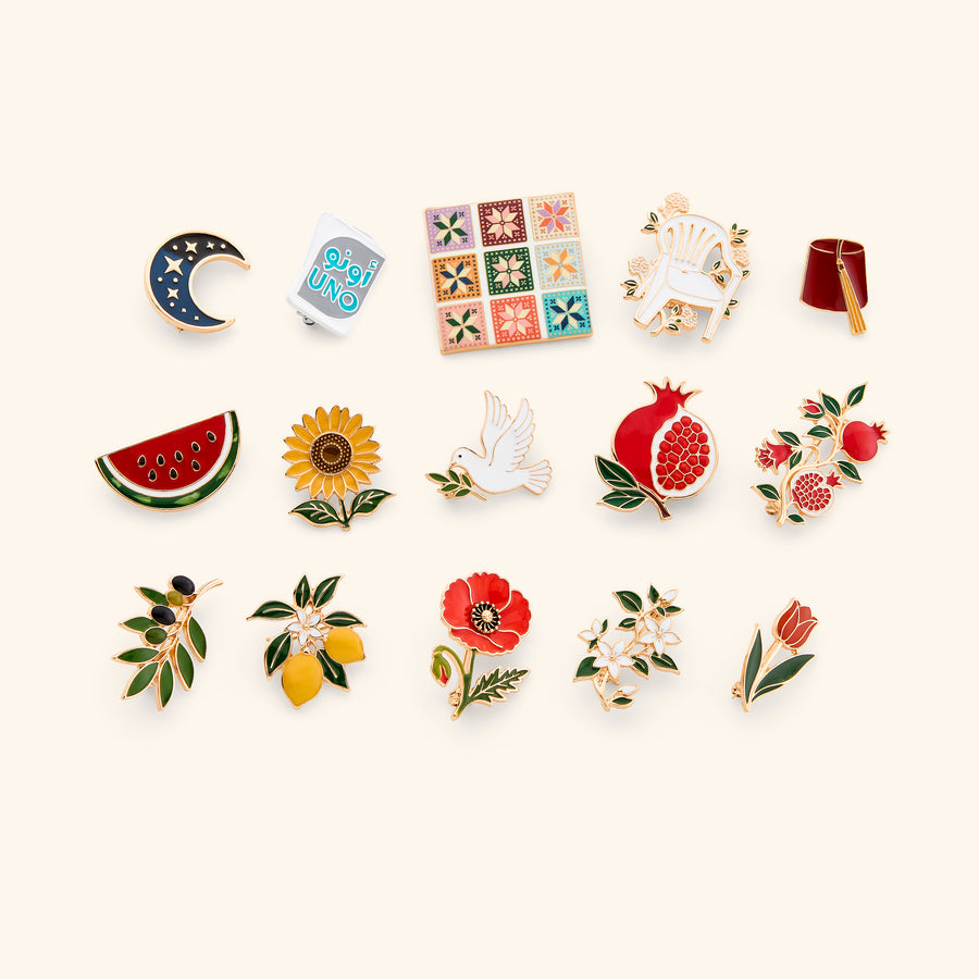 An assortment of enamel pins crafted with an 18k gold-plated finish featuring cultural and heritage motifs including a watermelon, olive branch, pomegranate, Tatreez tile, and Arabic Uno, displayed on an off-white background.