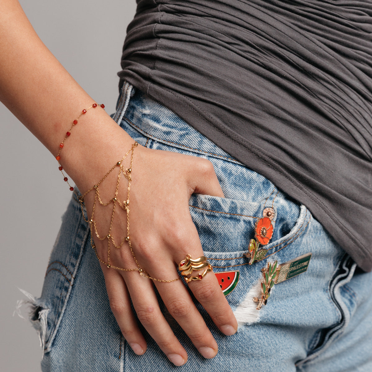On-model lifestyle view of the Essential Red Agate Micro Bracelet, featuring a draped hand chain and stacked ring, all in 18k gold-plated finish, resting on a denim jeans pocket.