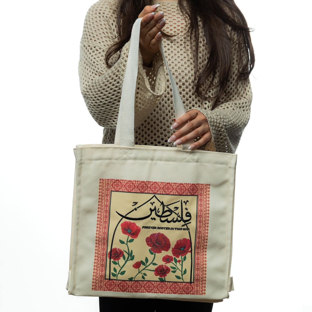 Woman in a beige textured sweater holding the Poppy Tatreez Palestine tote by the handles.