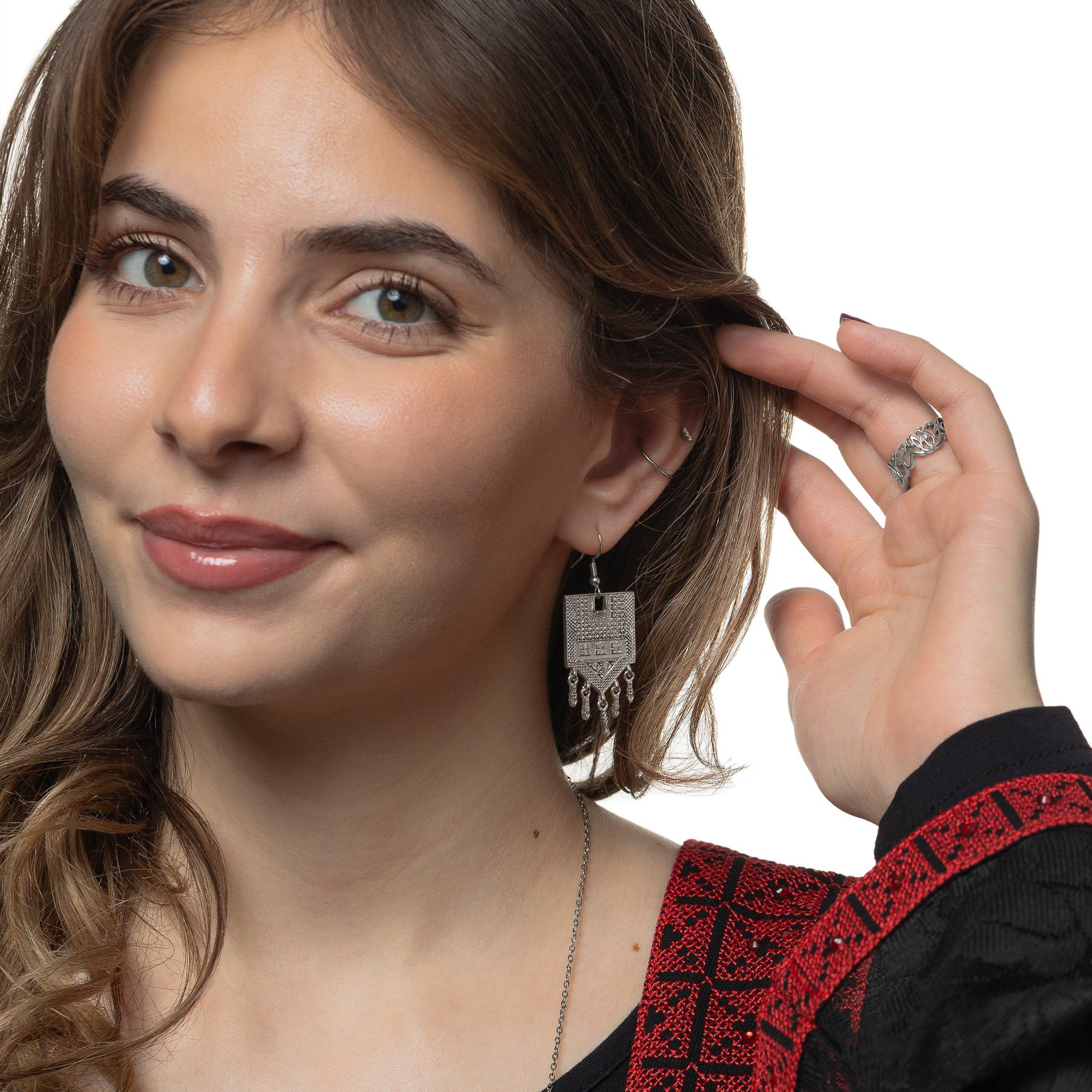 Photo of the model smiling to the camera, holding their hair away from their left ear. The model is wearing the silver Raining Tatreez Earrings