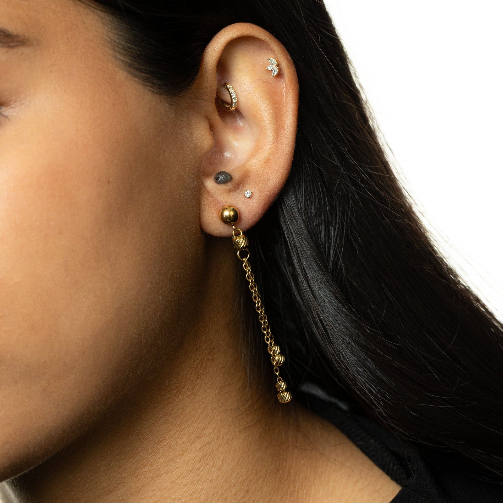 Woman wearing a single gold earring with double chain dangles ending in ribbed bead charms, styled with black hair tucked back.