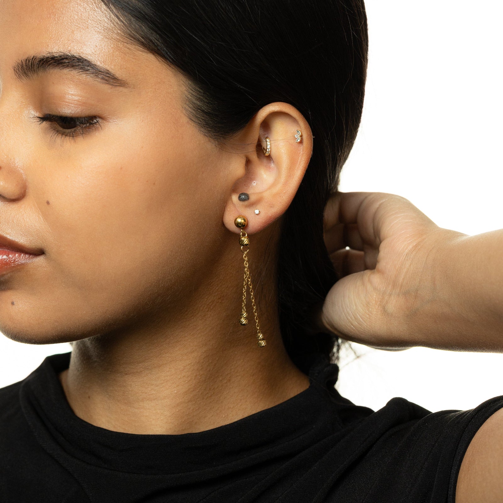 Woman in side profile wearing gold drop earrings with dual chains and ribbed beads, hand resting near the back of her head.