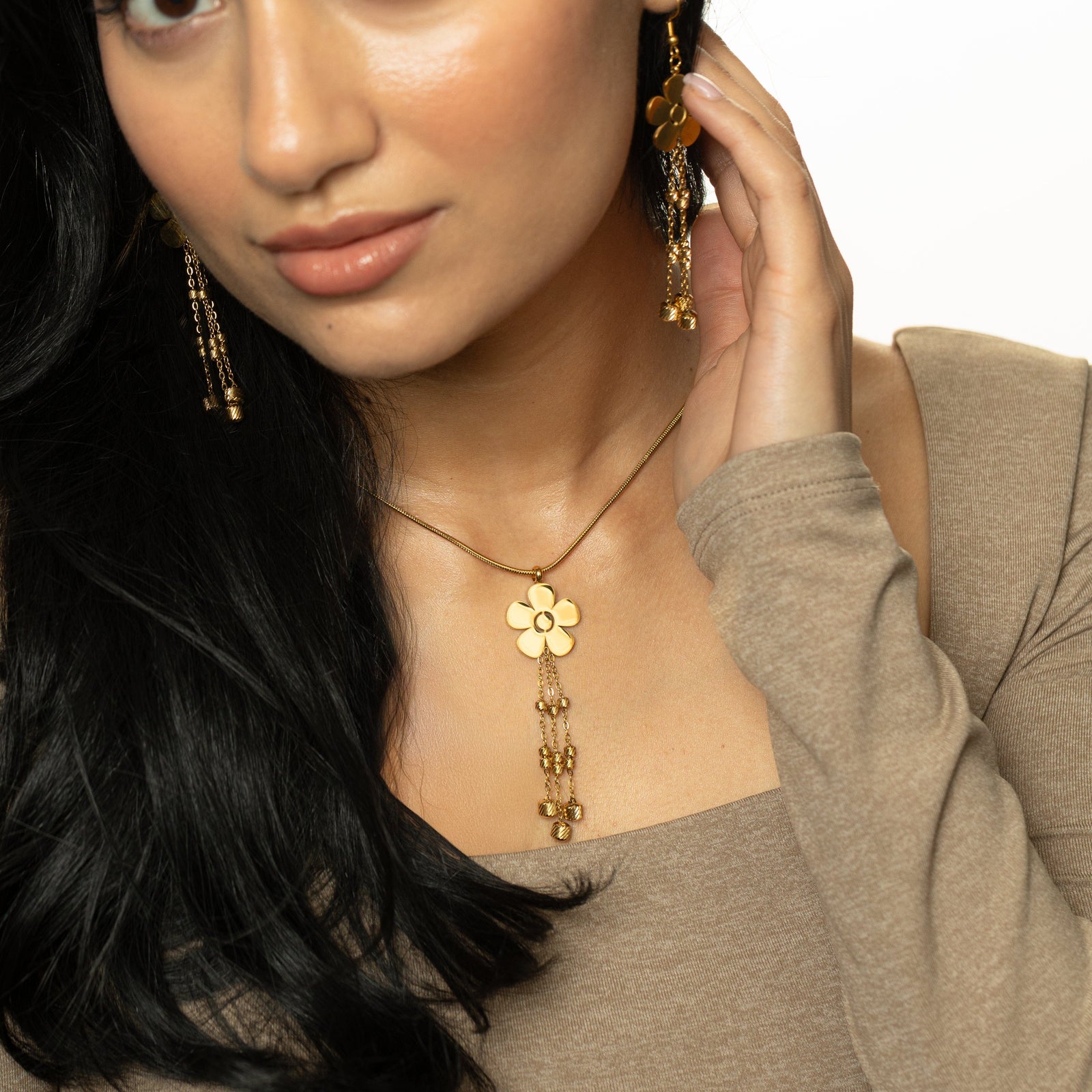 Detail shot of the gold Raining Flower Necklace pendant on the chest, styled with matching earrings, hand gently touching near the neckline.