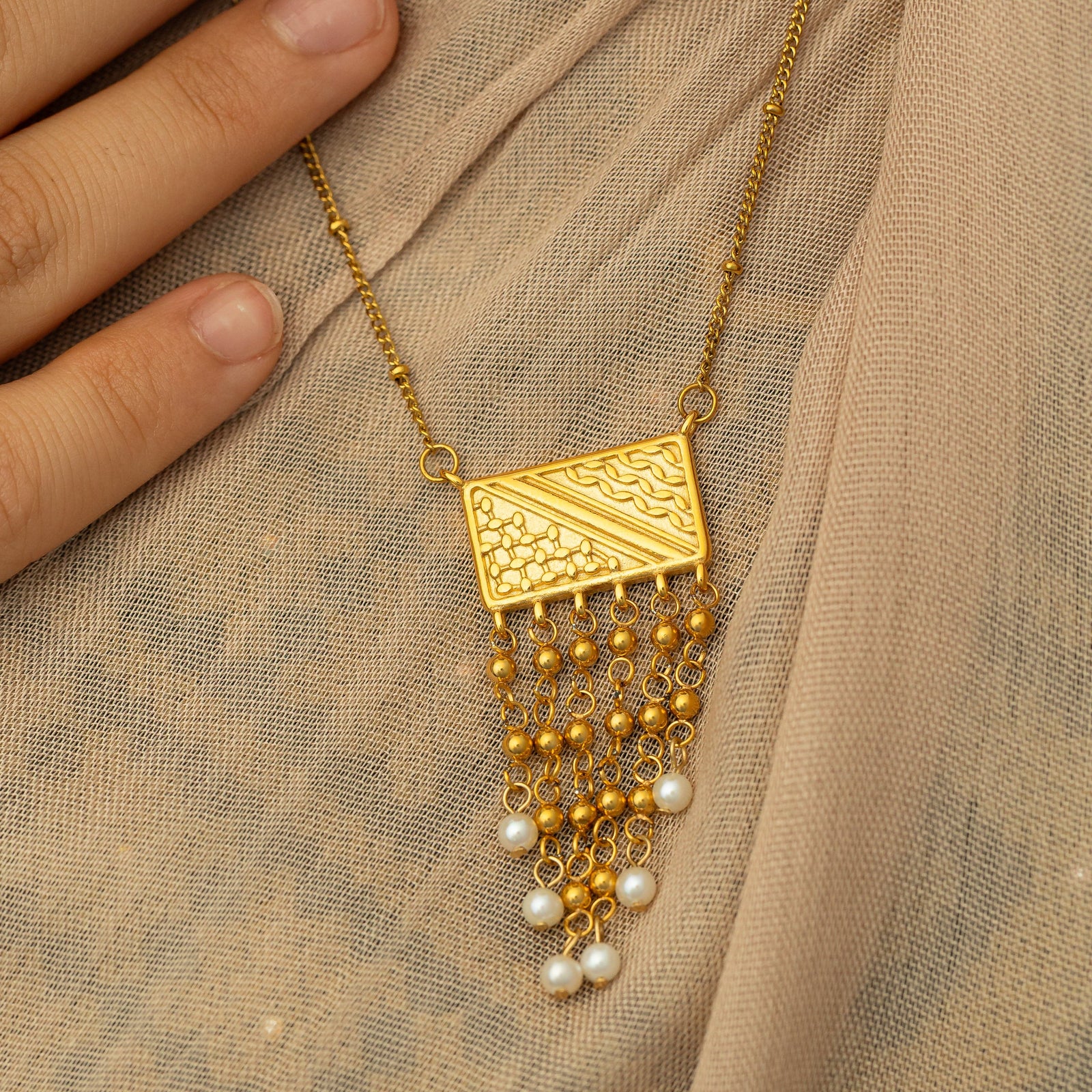 Close up photo of the pendant of the gold Raining Pearls Keffiyeh Necklace laying on a fabric. Model is touching the gold chain.