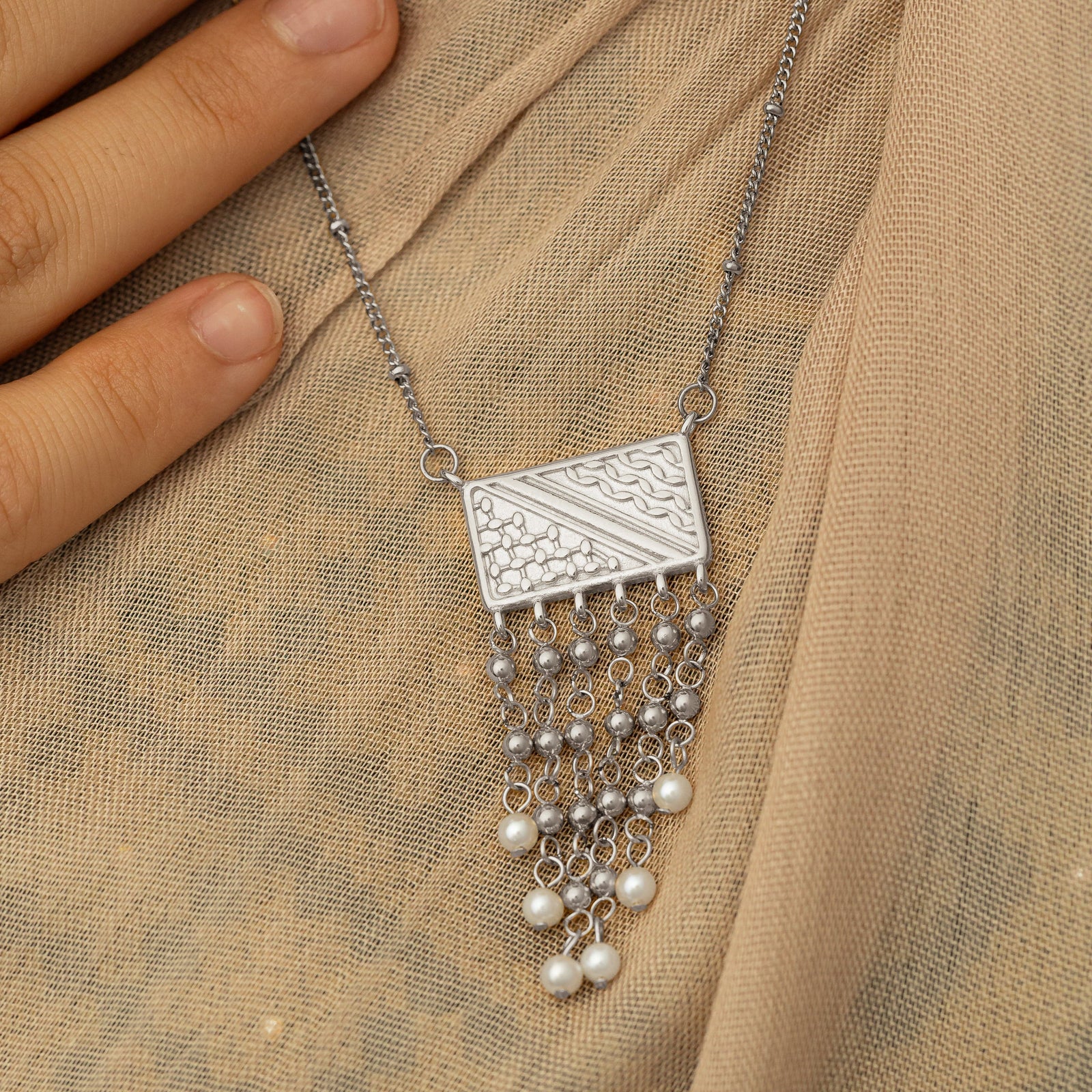 Close up photo of the pendant of the silver Raining Pearls Keffiyeh Necklace laying on a fabric. Model is touching the silver chain.